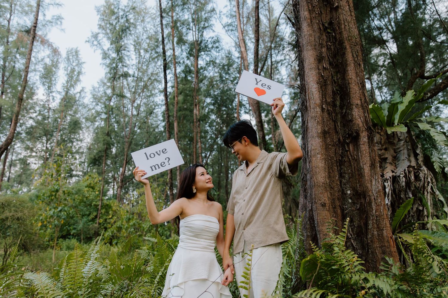 Couple in forest holding love signs during romantic outdoor engagement photo shoot.