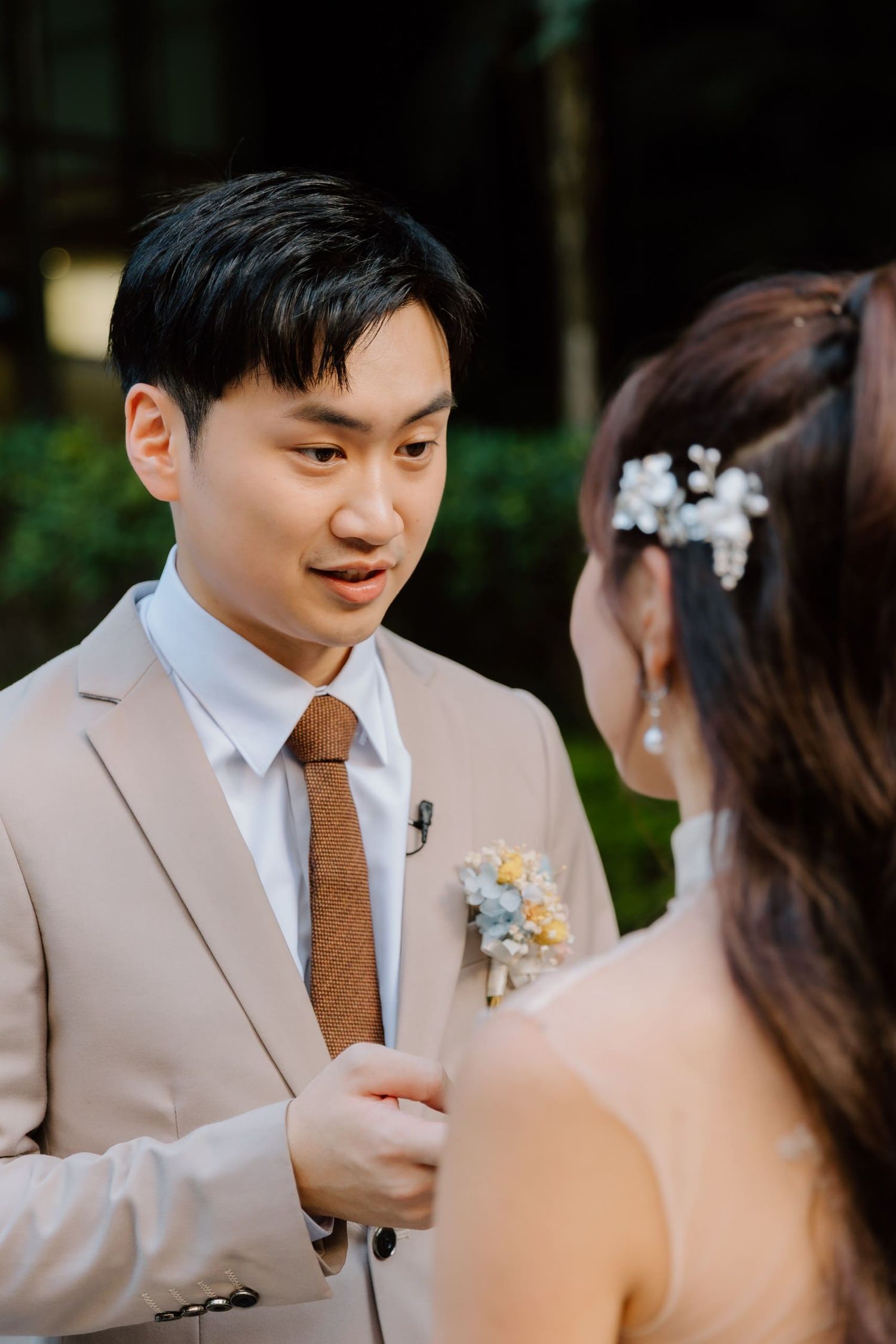 A groom in a beige suit with a brown tie holds his bride's hands and gazes at her tenderly during their wedding ceremony.