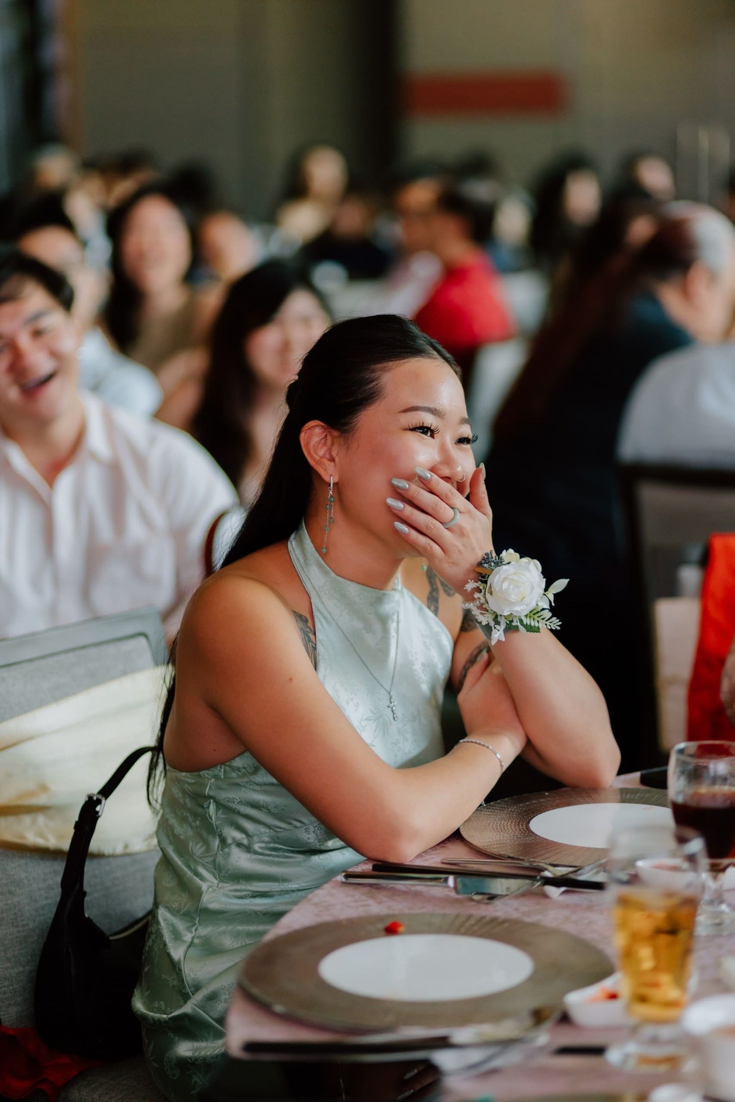 A joyful woman in a sage green dress with a floral wrist corsage laughs at a wedding reception dinner.