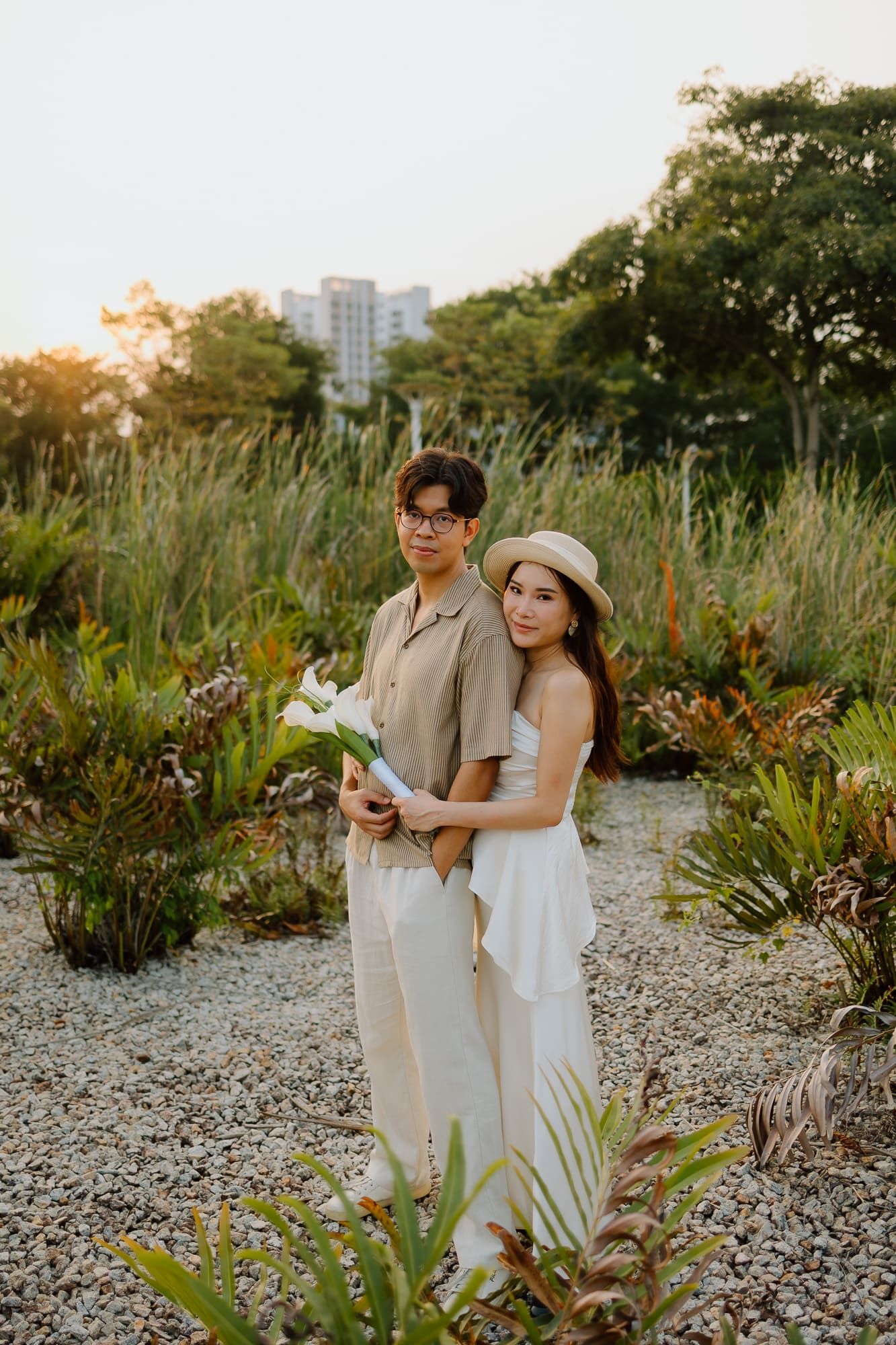 A couple poses in a lush garden at sunset, woman in white dress and hat holding flowers, man in beige outfit beside her.