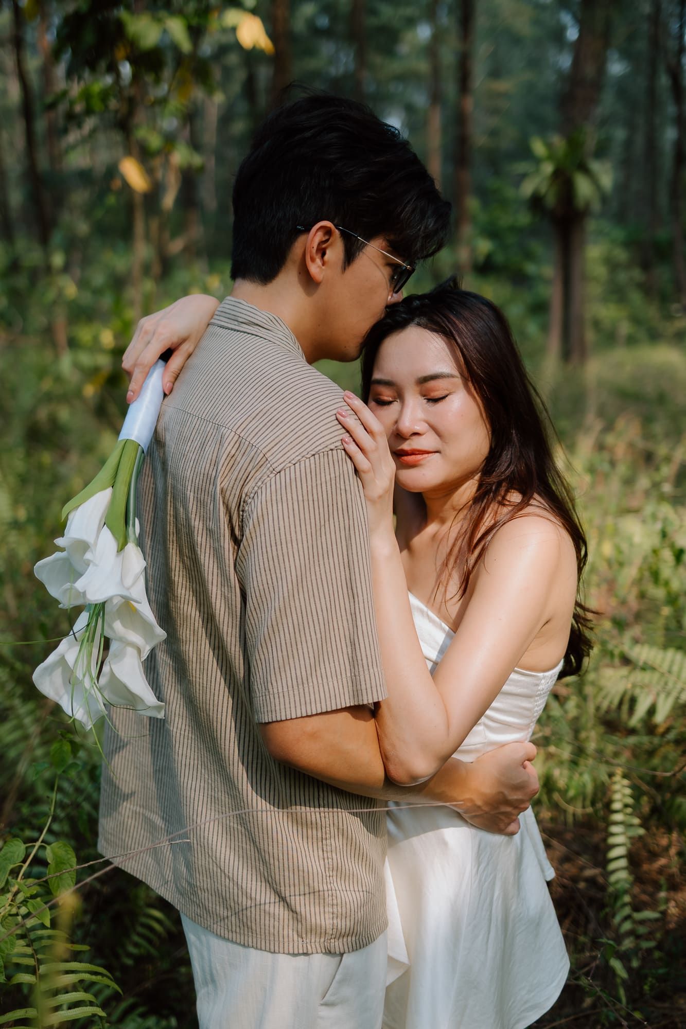 A couple embraces tenderly in a lush green forest, woman in white dress holds man carrying white flowers.