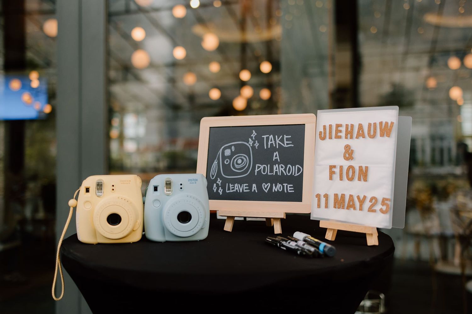Polaroid Instax Mini cameras and small chalkboard signs displayed at a wedding reception with bokeh lights in background.