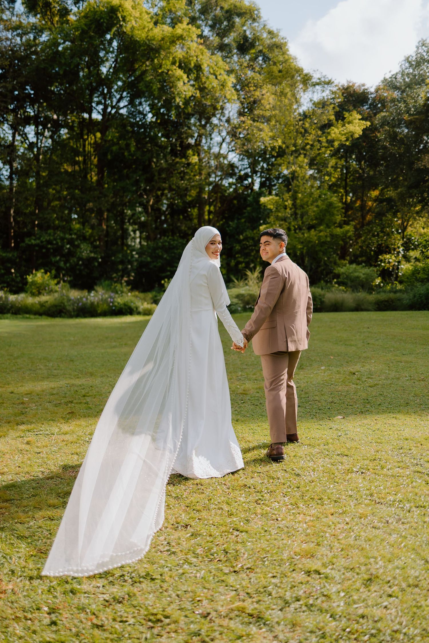 Muslim bride in white hijab and gown holds hands with groom in tan suit, walking in lush green park.