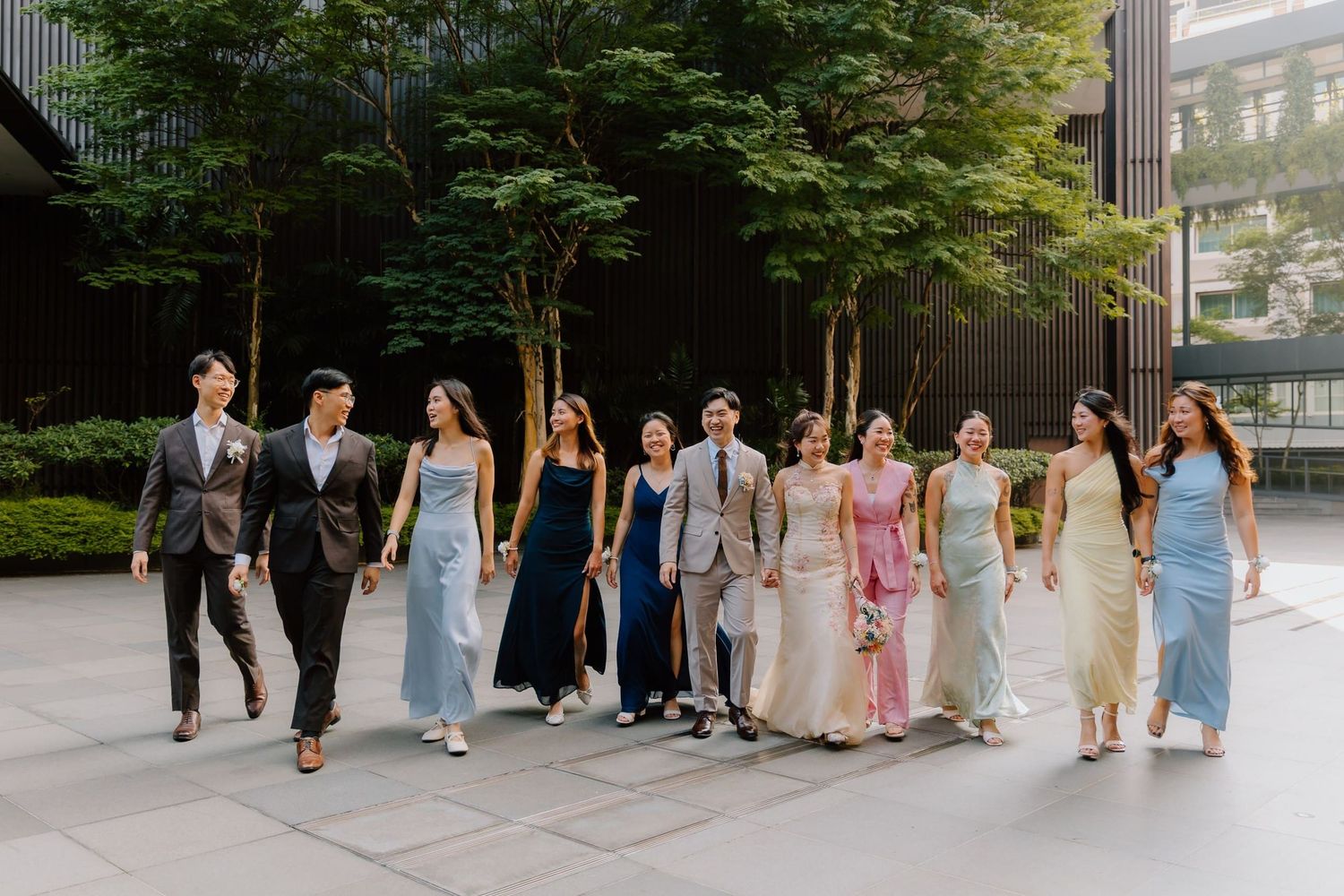 A stylish group of young adults in elegant formal attire walk together outdoors near modern buildings and lush trees.