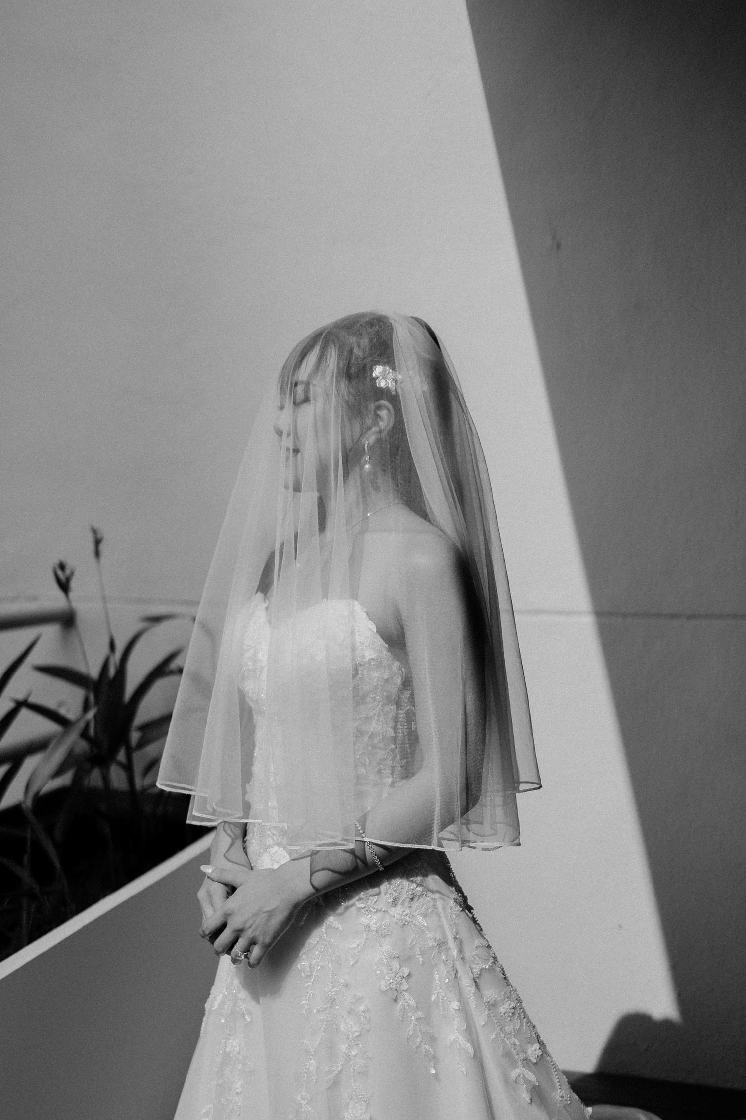 Black and white photo of a bride in a strapless lace gown and veil, standing in soft light against a white wall.