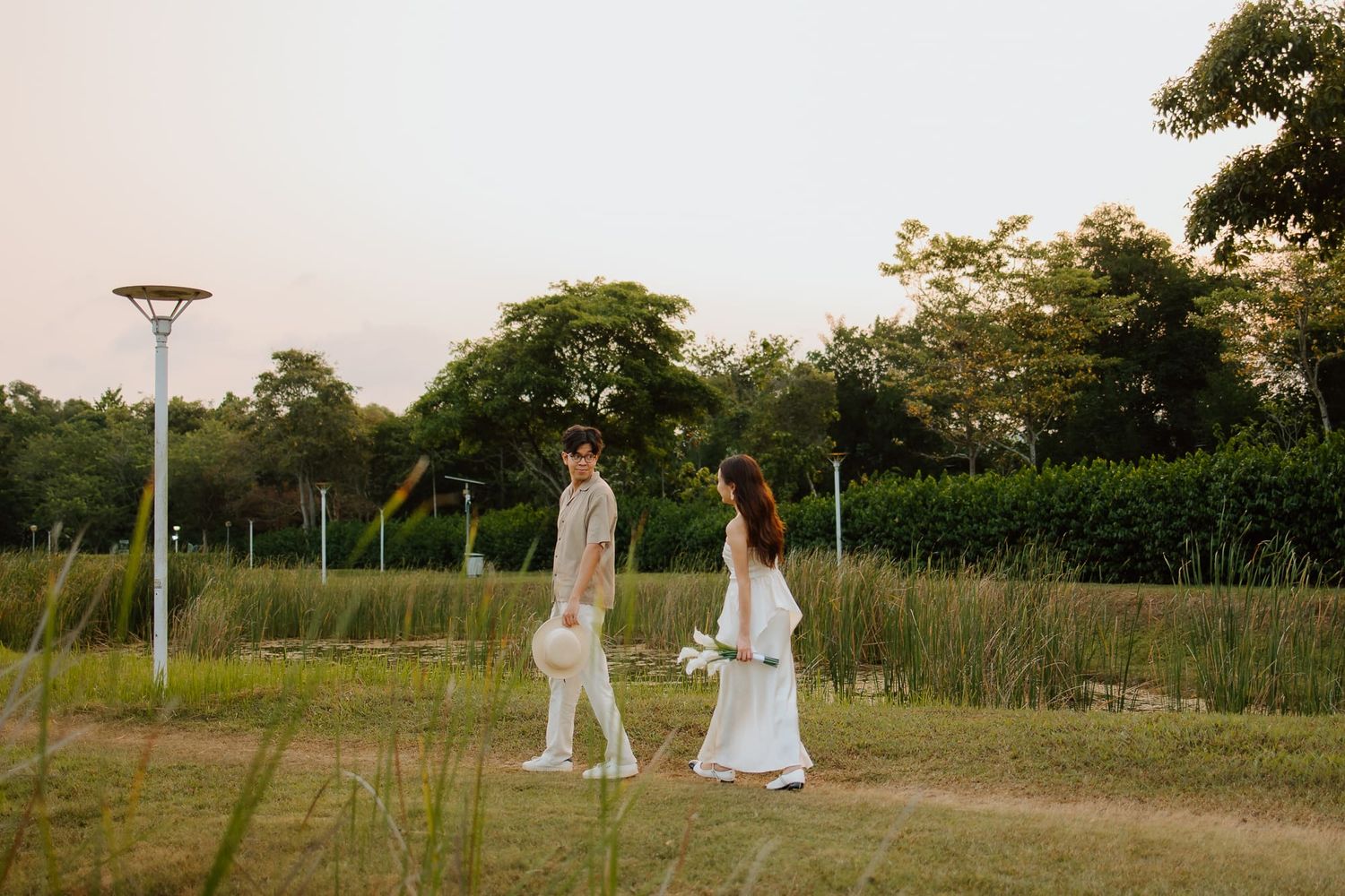 A bride and groom walk through a lush green park at dusk, the bride holding a white bouquet in an elegant gown.