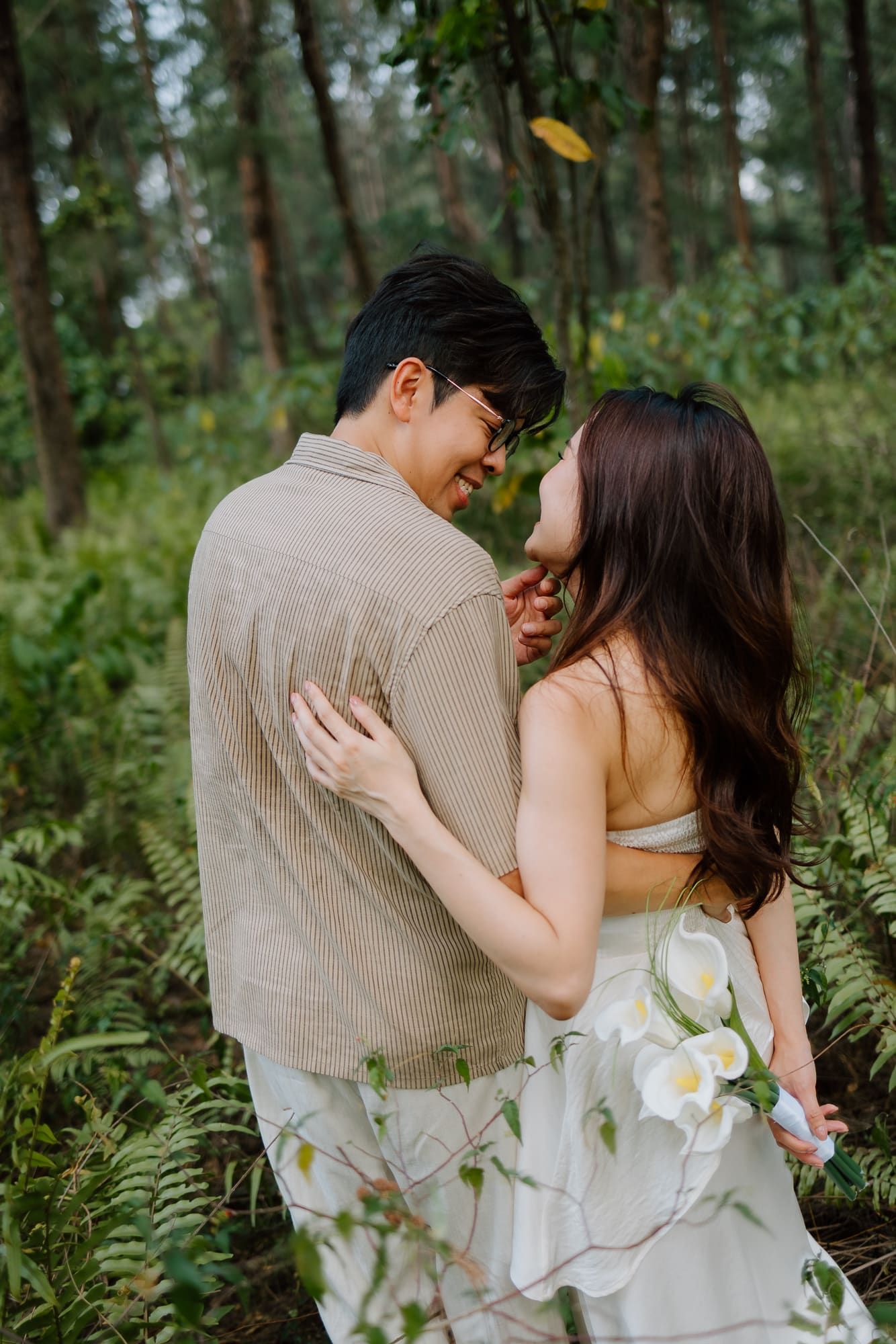 A couple embraces romantically in a lush forest, woman in white dress holding flowers, man in beige shirt smiling.
