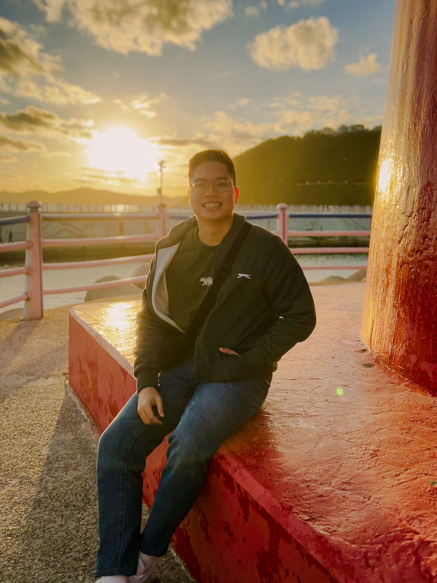 Man in black jacket sitting on red wall at sunset with scenic waterfront and mountains in background.