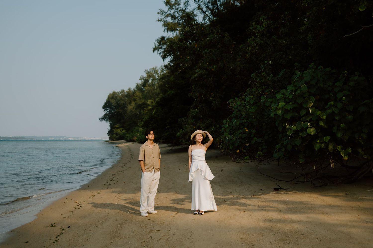 Couple walking on a serene tropical beach with lush green trees, man in beige and woman in white dress.