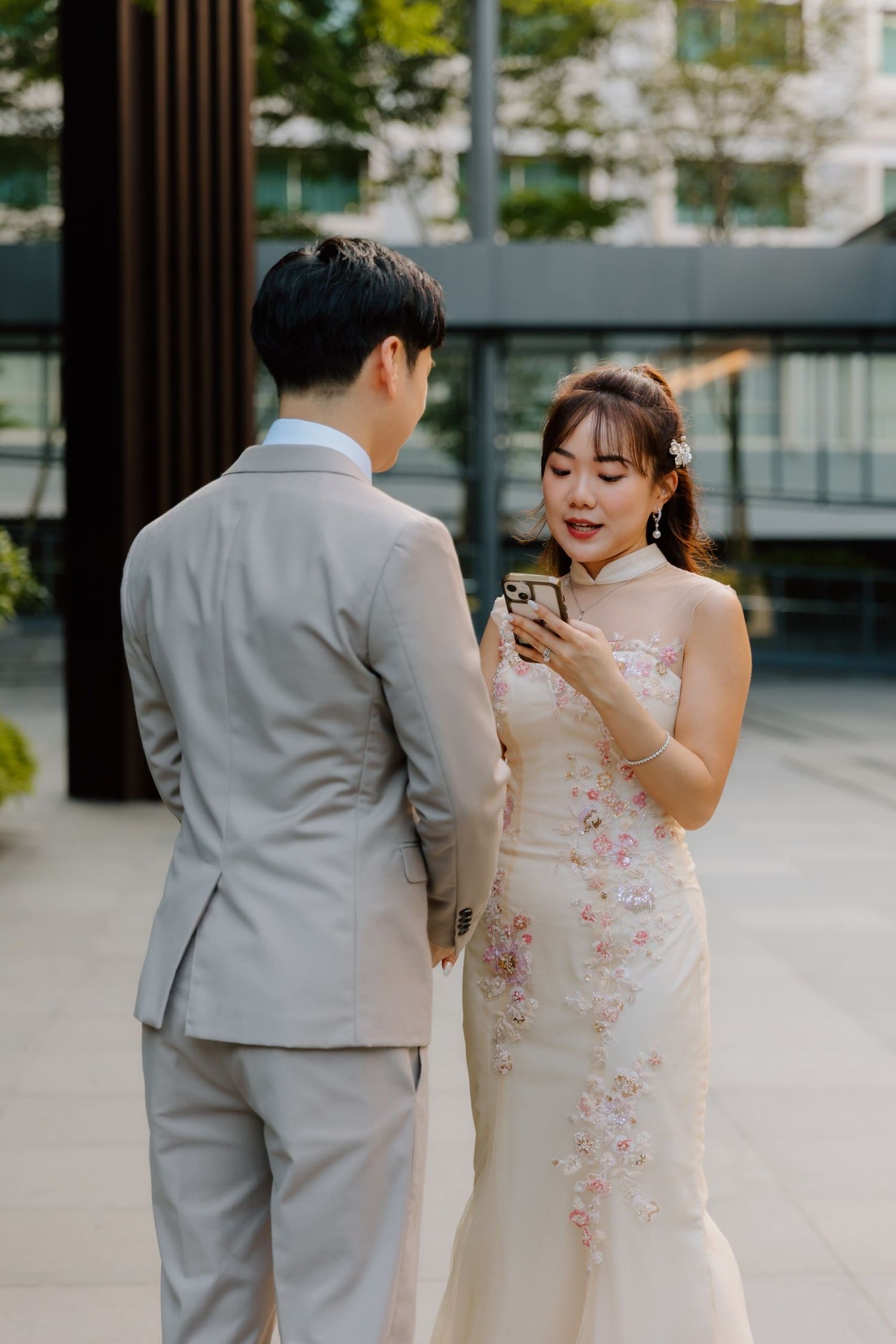 Bride in floral qipao reads vows on phone beside groom in gray suit at outdoor wedding venue.