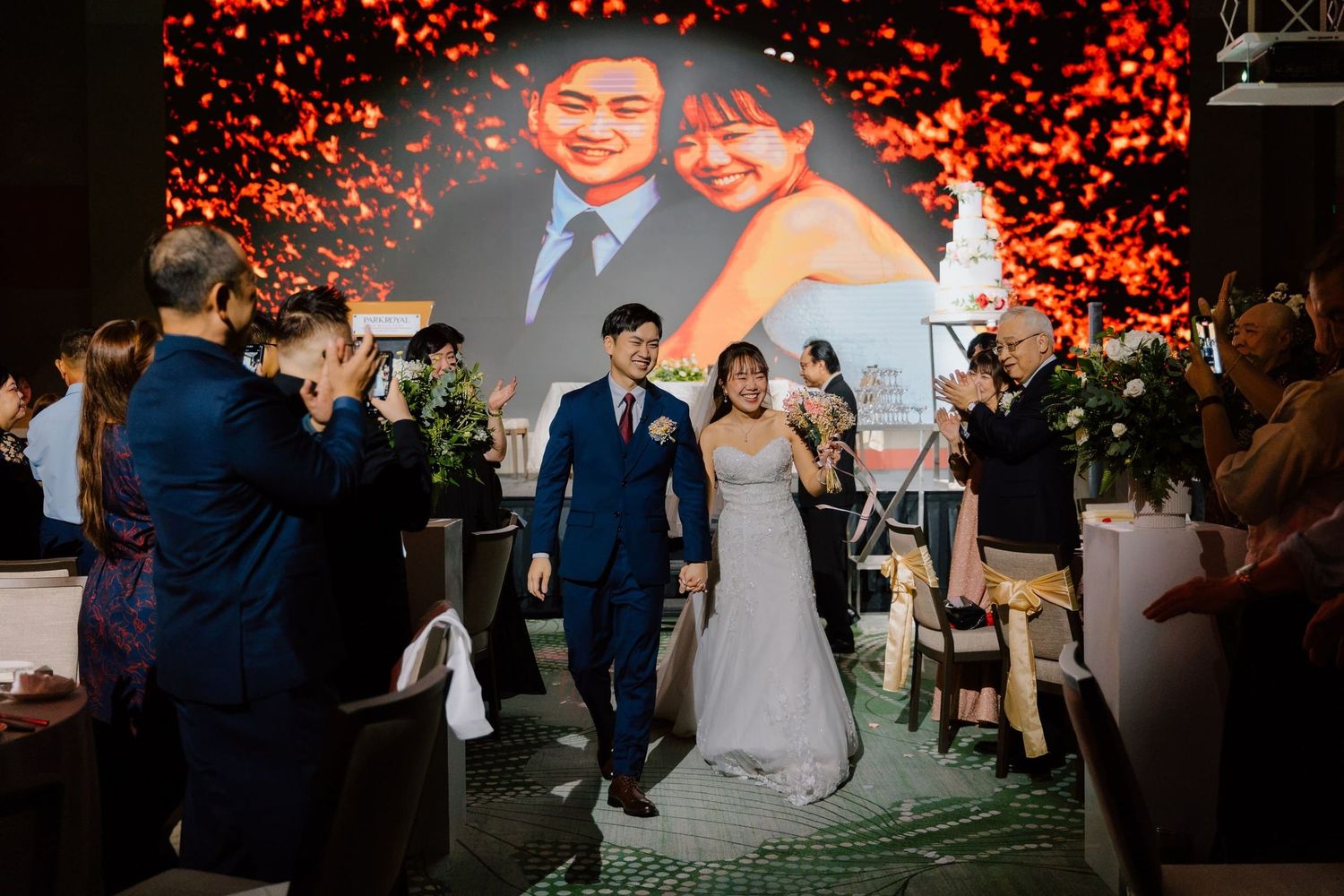 A couple walks down the aisle at their wedding reception with a romantic photo displayed on a large screen behind them.