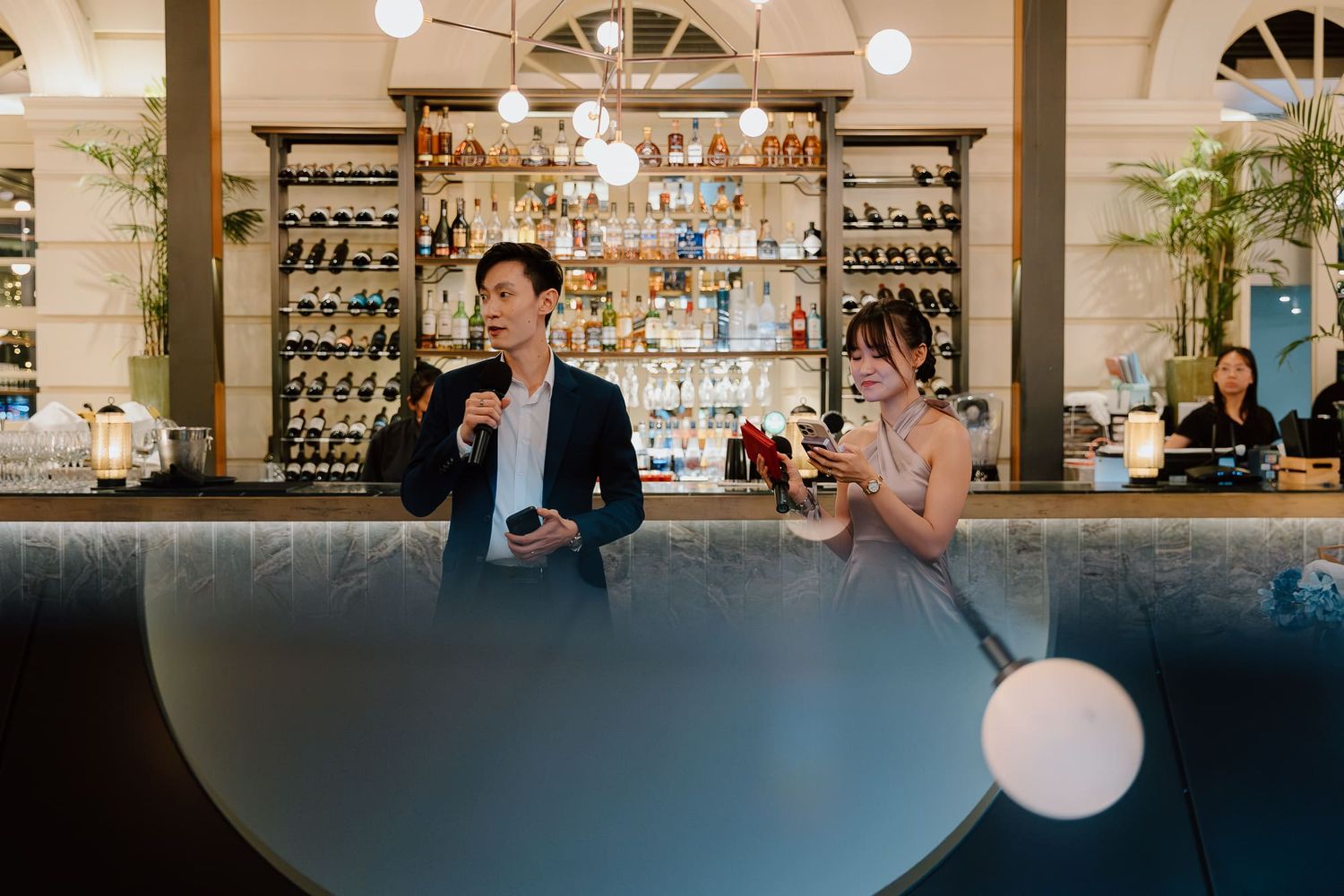 Restaurant patrons sit at a bar inside an elegant establishment with arched windows and pendant lighting.