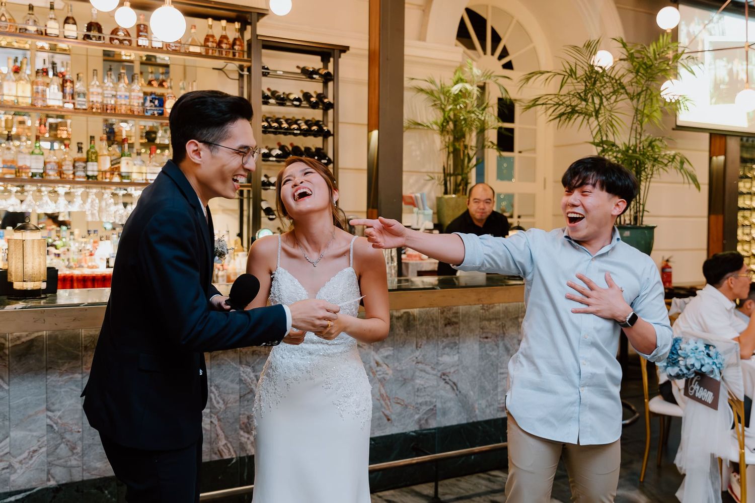 Wedding guests share a joyous laugh together at an elegant bar with illuminated shelves of liquor bottles.