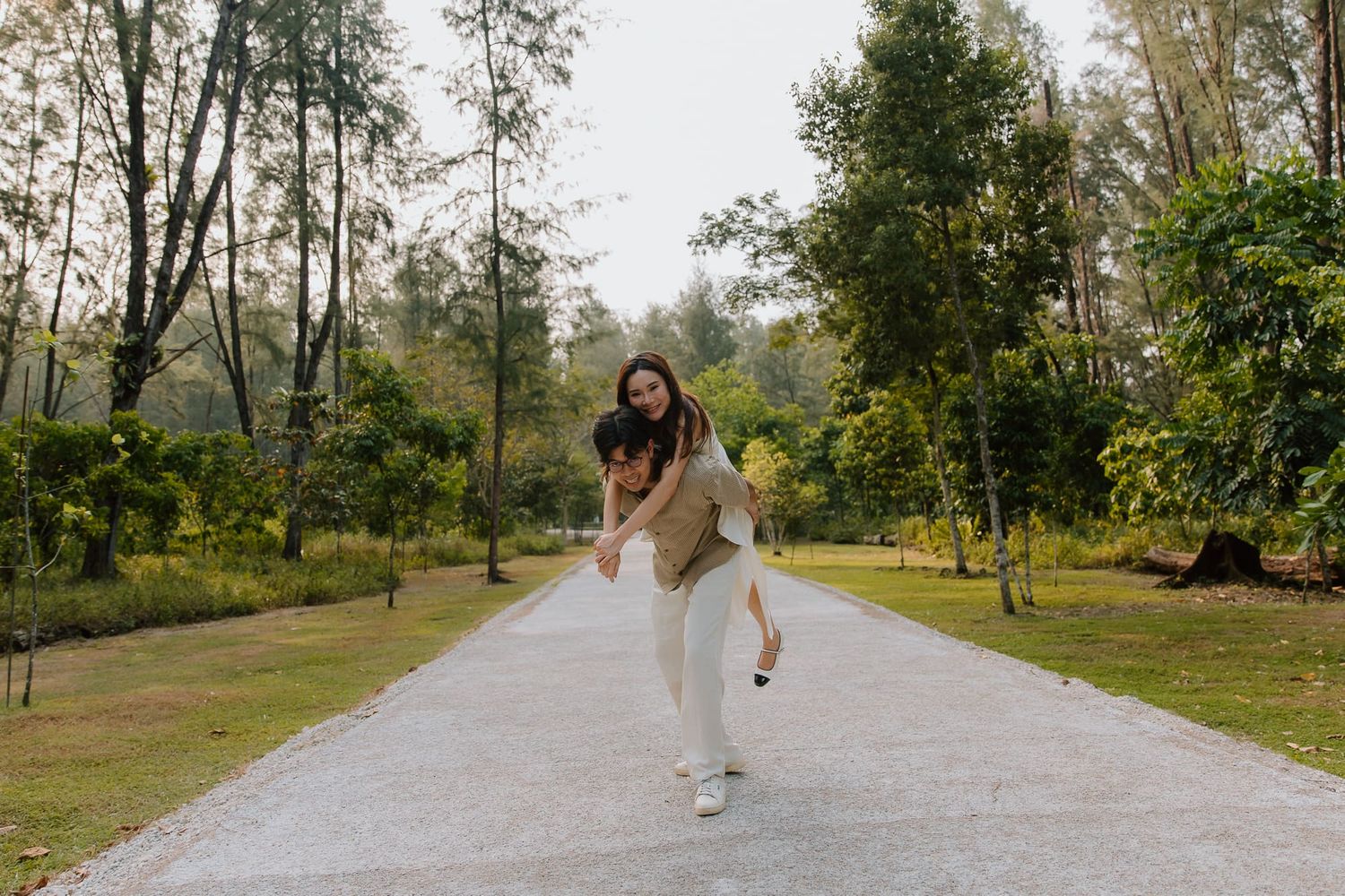 Couple enjoying a piggyback ride on a scenic park pathway surrounded by lush green trees.