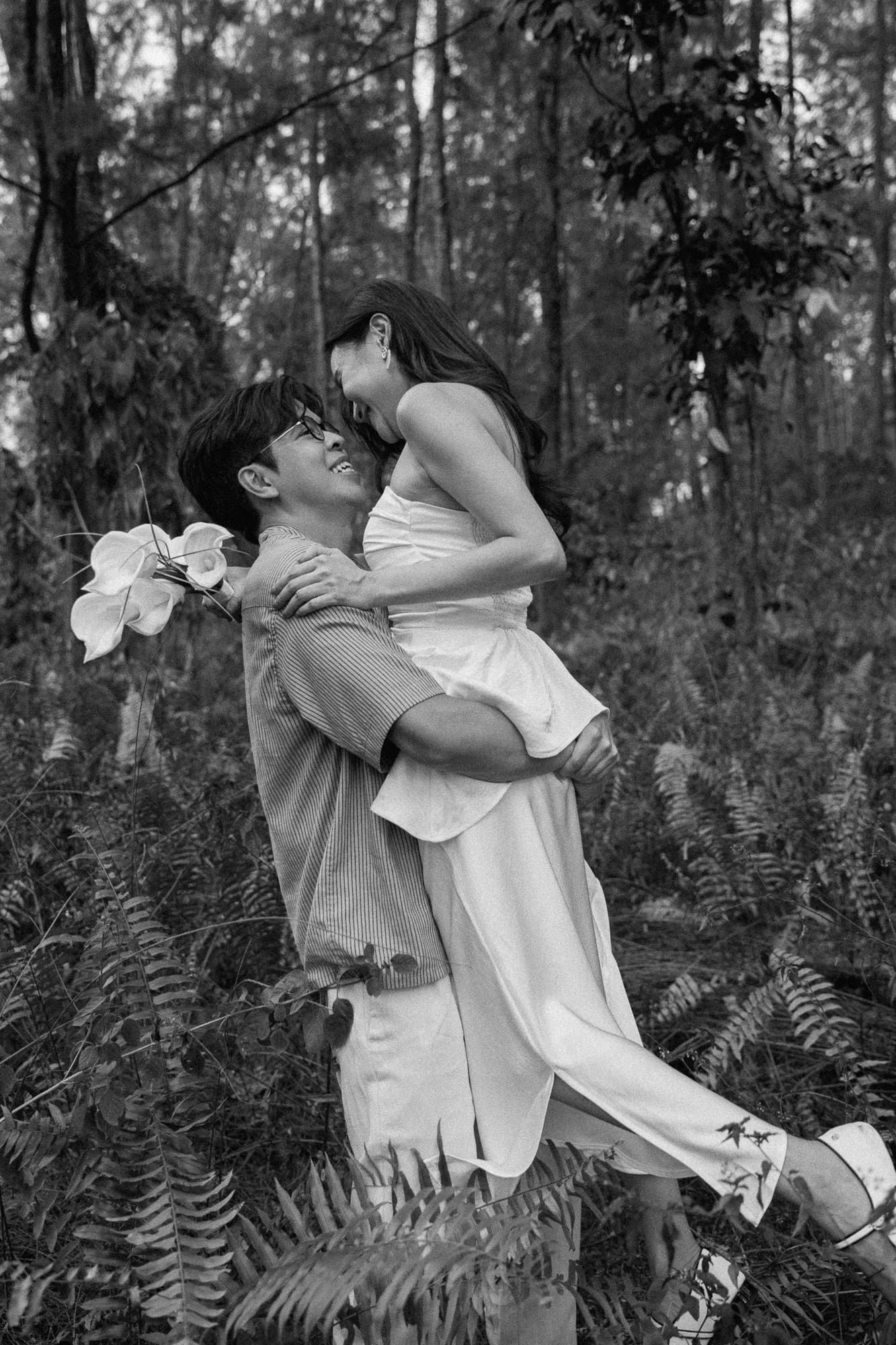 Black and white photo of a couple embracing in a forest, man lifting woman while she laughs, holding white flowers.