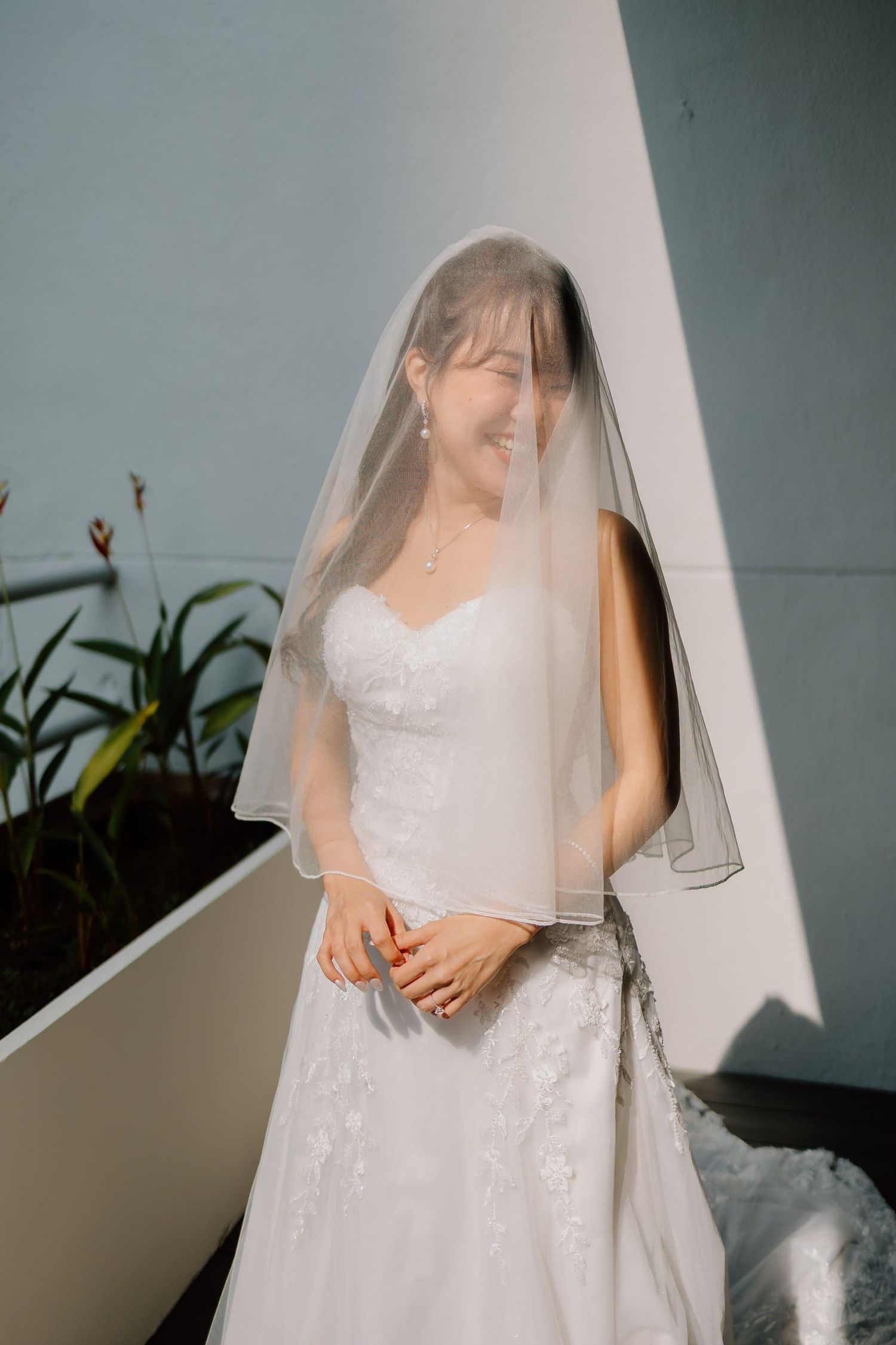 Smiling bride in white lace wedding dress and veil standing outdoors in soft sunlight.