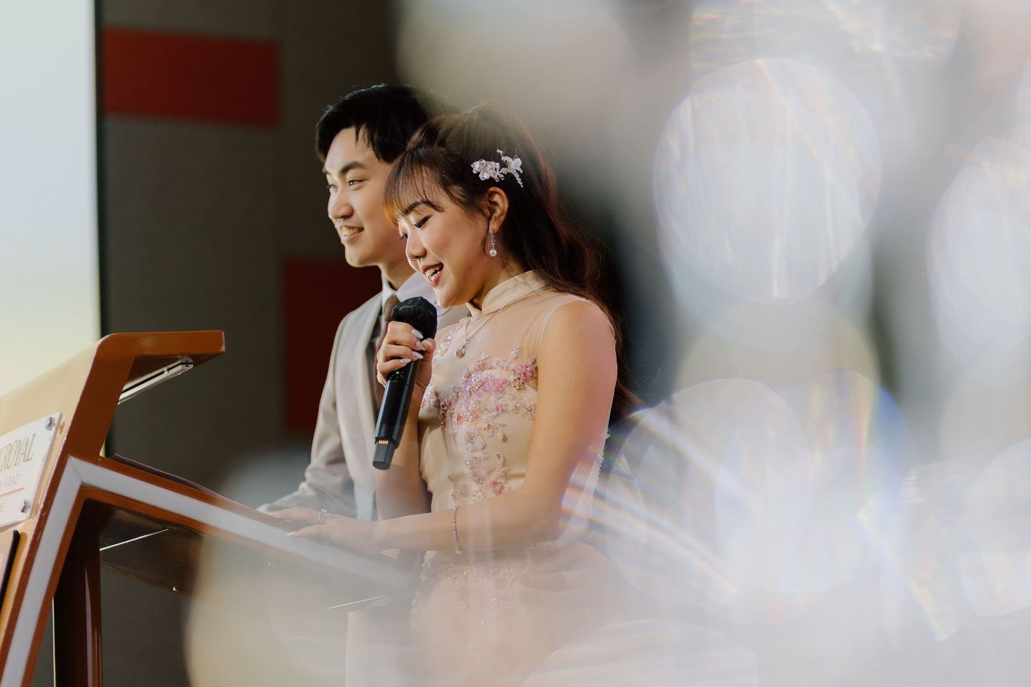 A couple shares a microphone at a wedding reception podium, the bride wearing a floral dress with hair accessories.