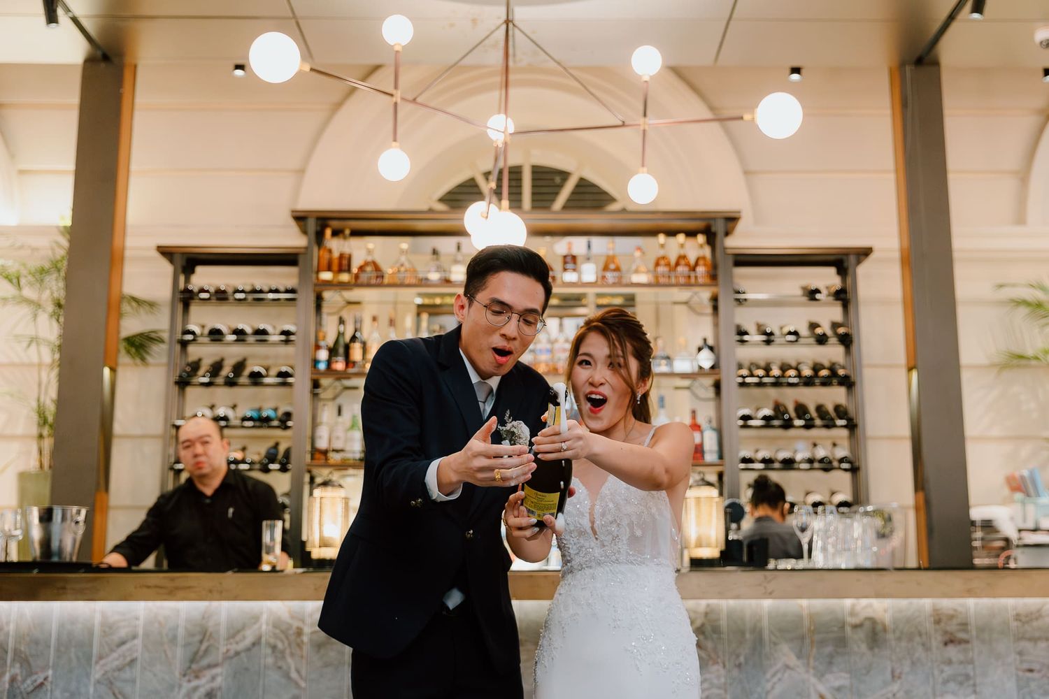 Two people stand at a stylishly lit cafe counter with pendant lights and glassware displaying in the background.