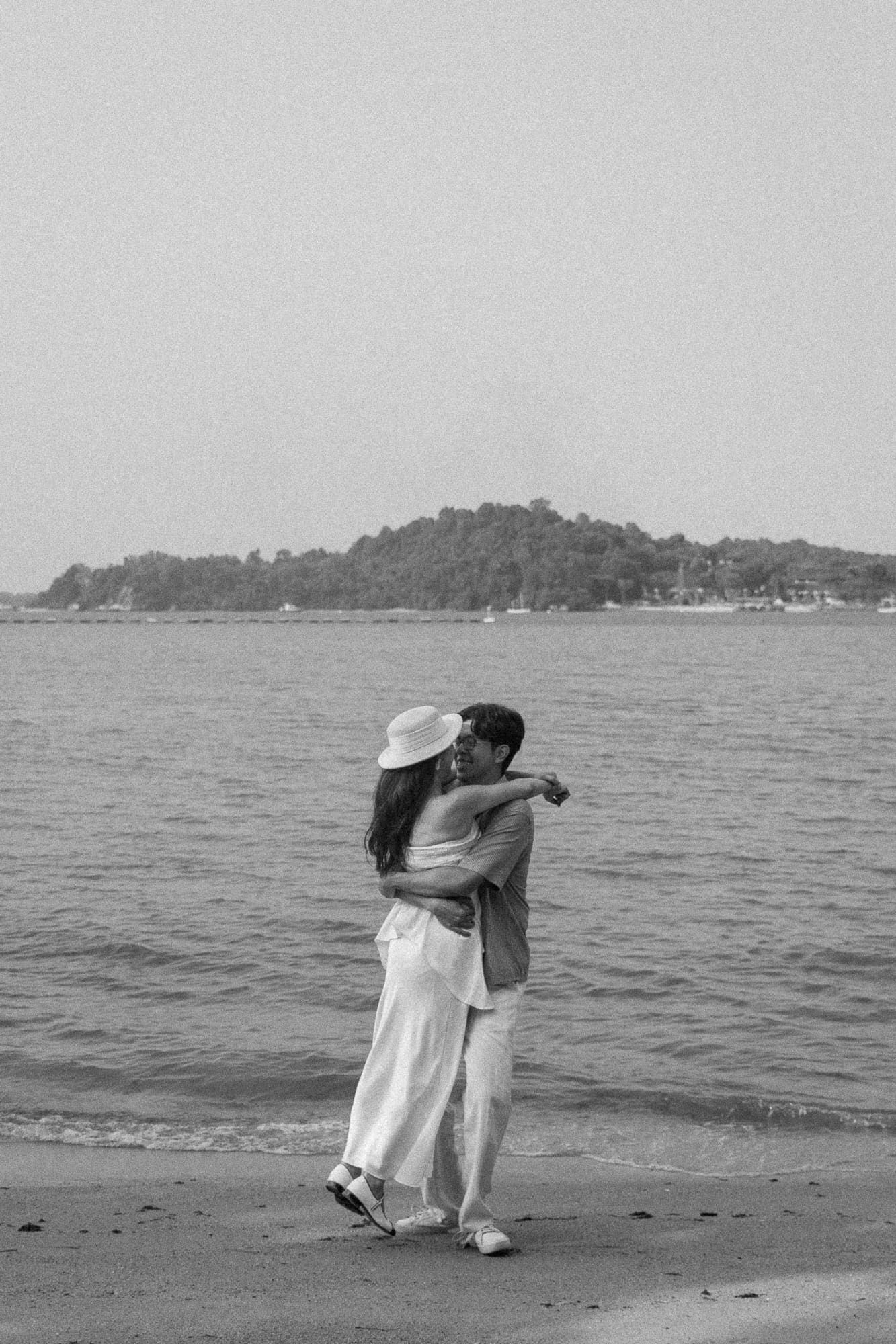 Black and white photo of a couple embracing on a beach with an island and calm ocean in the background.