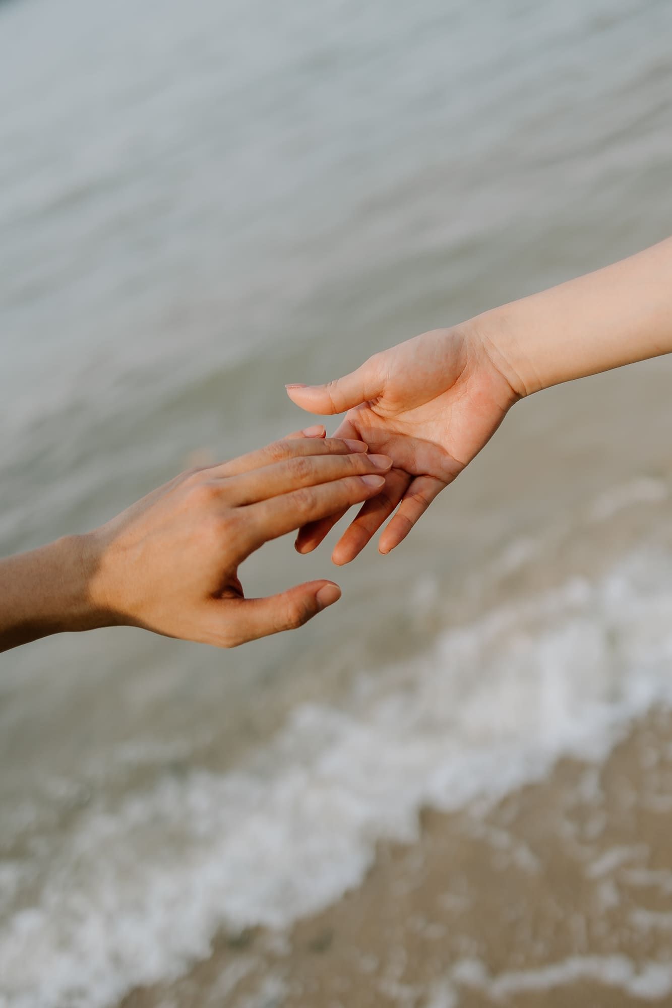 Two hands of different skin tones gently touching fingertips on a sandy beach with ocean waves in the background.