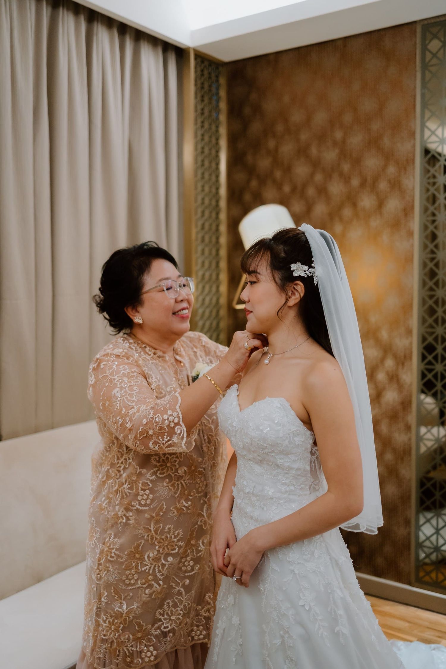 An older woman helps a bride put on a necklace as she prepares for her wedding day in a elegant room.