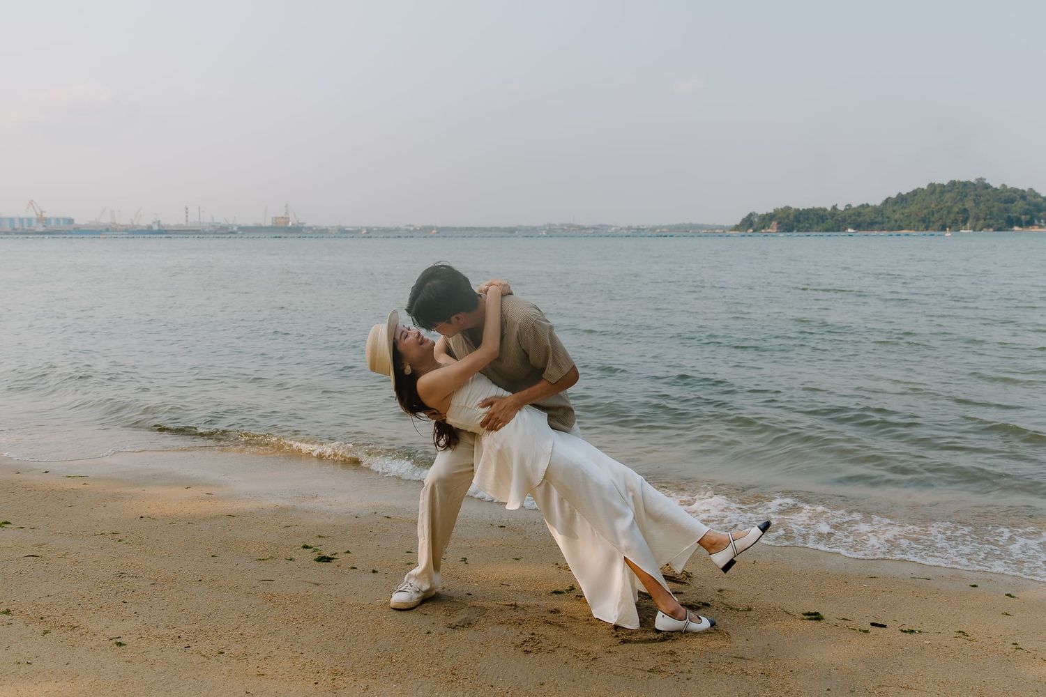 Couple sharing a romantic dip kiss on a sandy beach shore with calm ocean waters and green hills in the background.