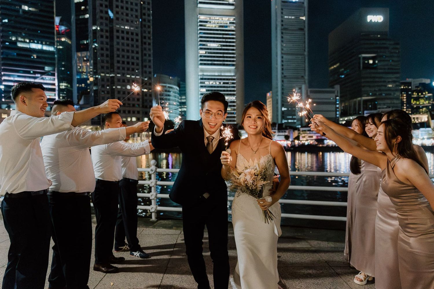 A wedding party takes photos at night with illuminated city buildings in the background creating a romantic urban setting.