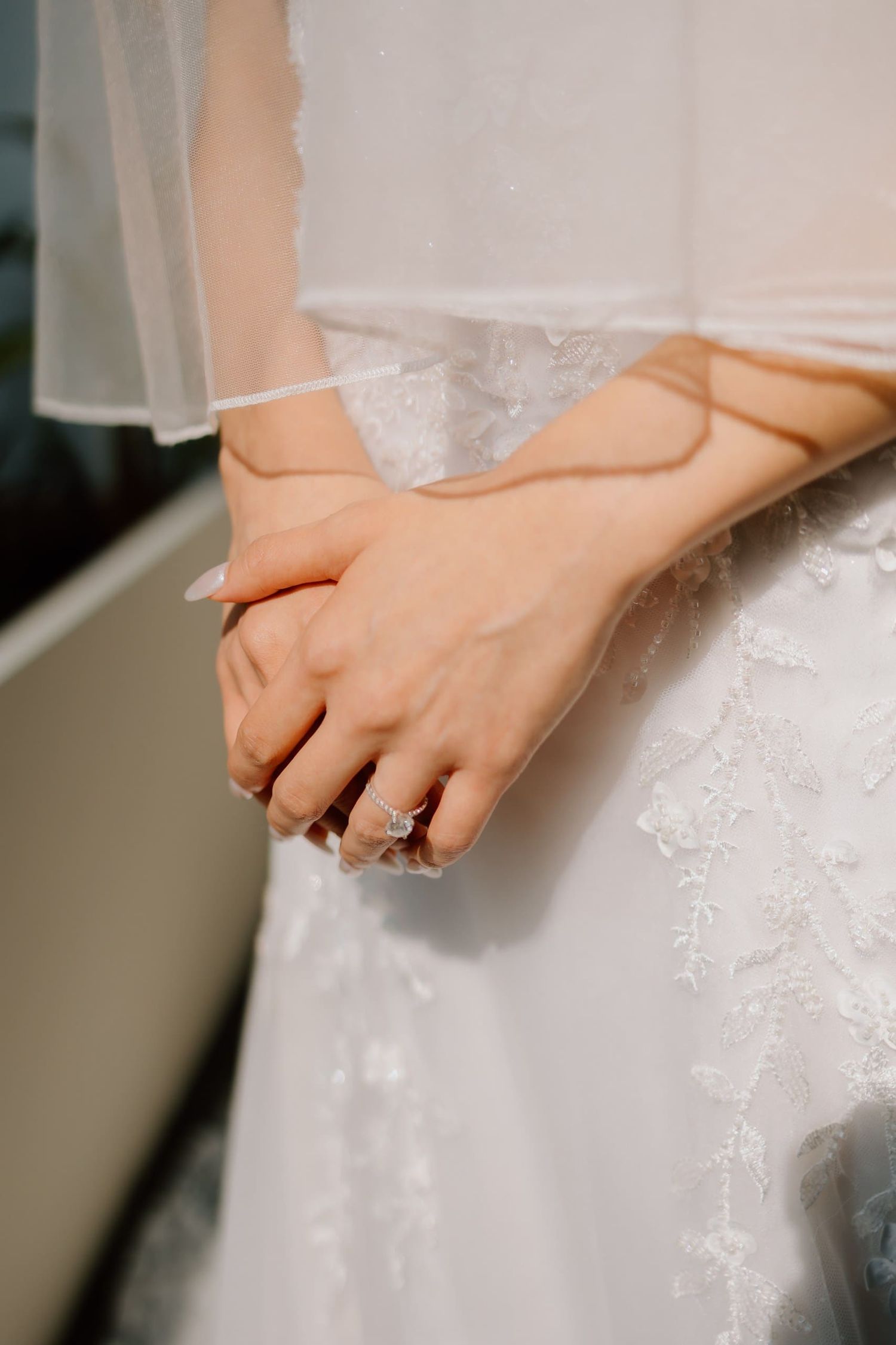 Bride in white lace gown clasping hands, showcasing a sparkling diamond engagement ring with veil detail.