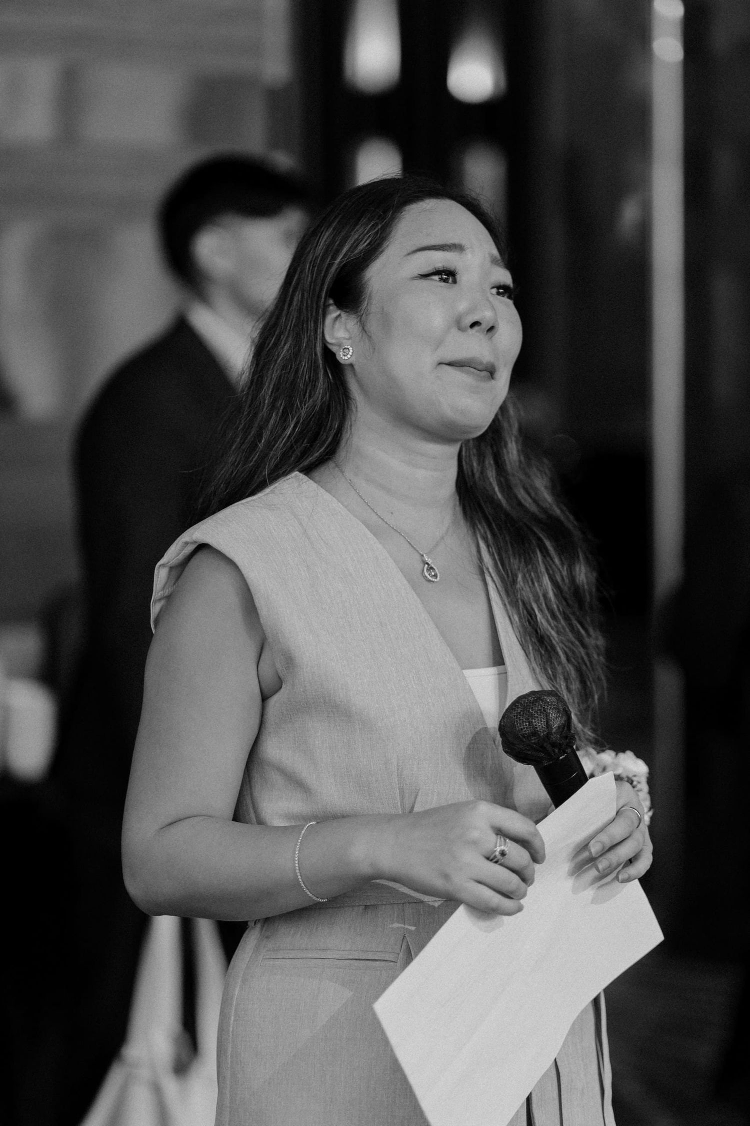 Black and white photo of a woman holding a microphone and papers, speaking at a formal event.