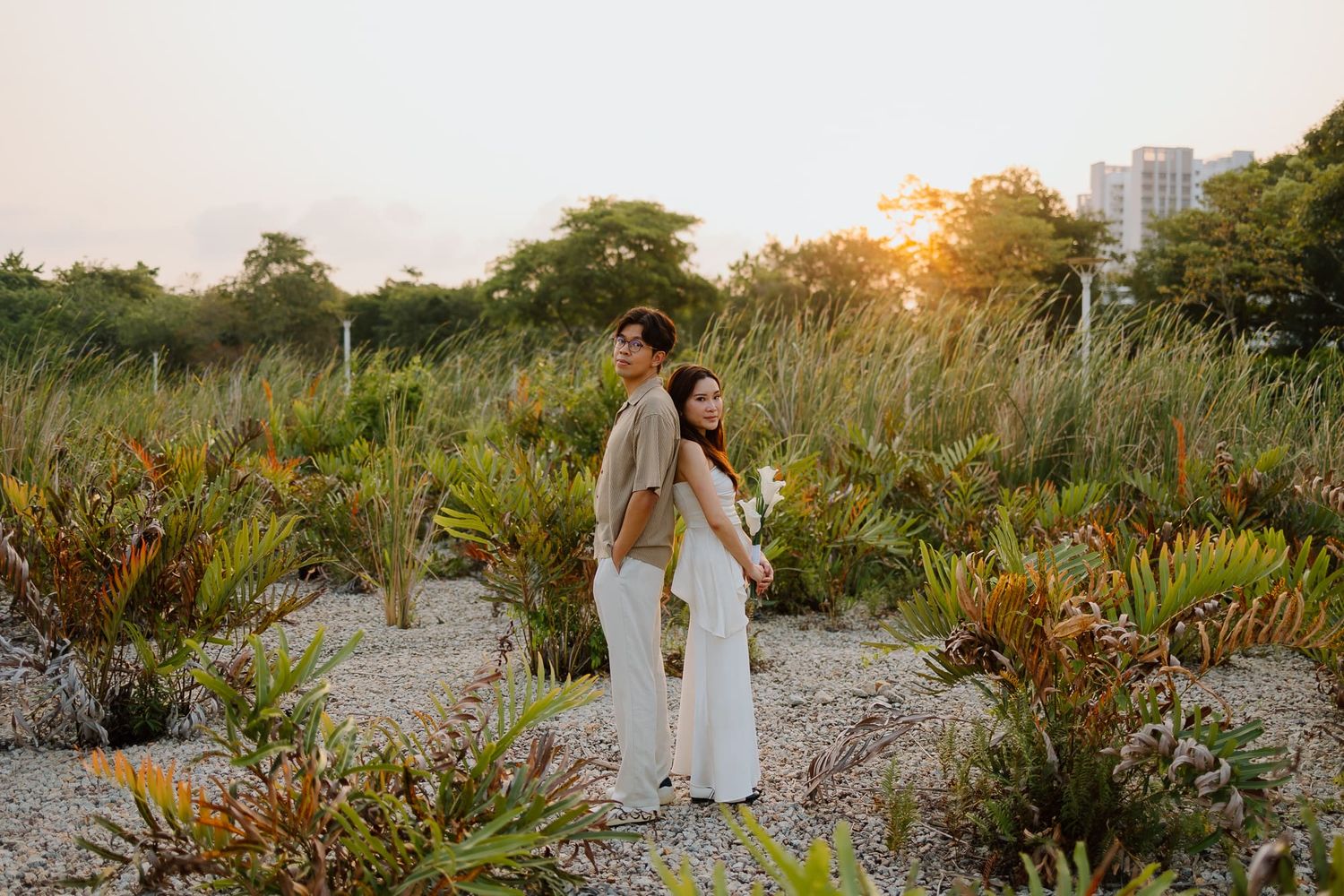 Couple standing back-to-back in a lush tropical garden at golden sunset with city buildings in the background.
