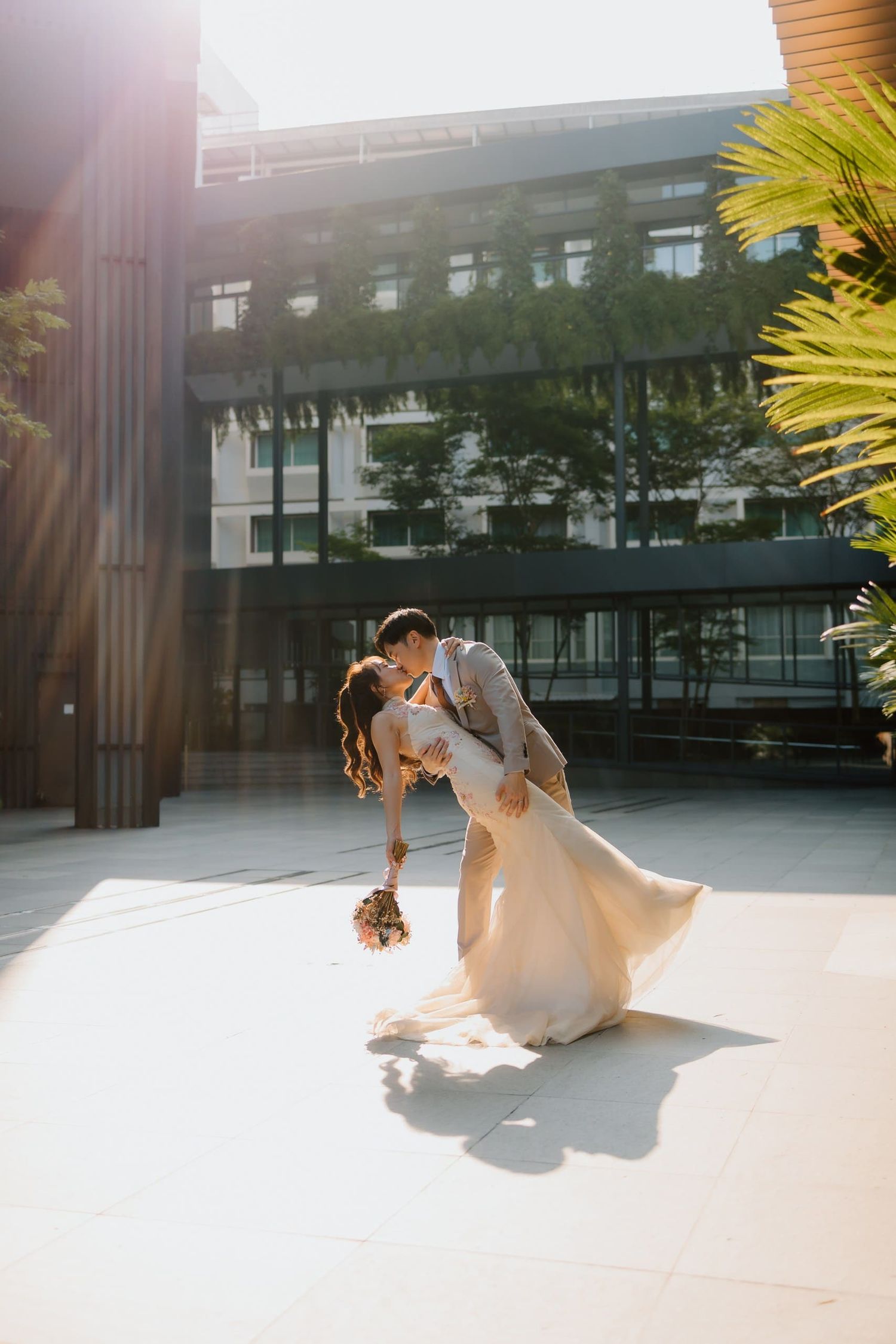 A couple shares a romantic dip kiss in golden sunlight, bride in flowing gown holding bouquet, groom in gray suit outdoors.