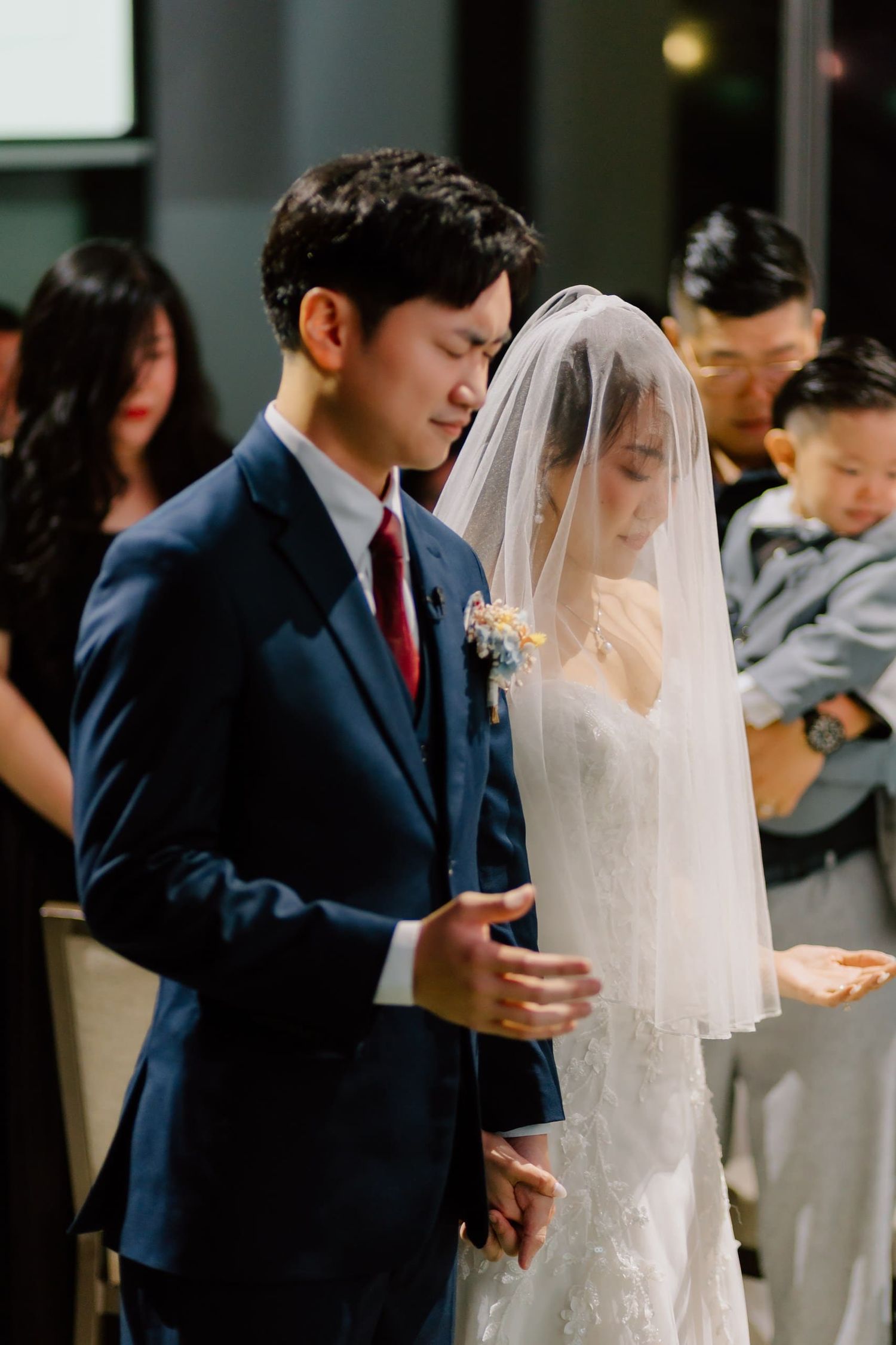 A groom in a navy suit and bride in a white lace gown with veil pray together during their wedding ceremony.