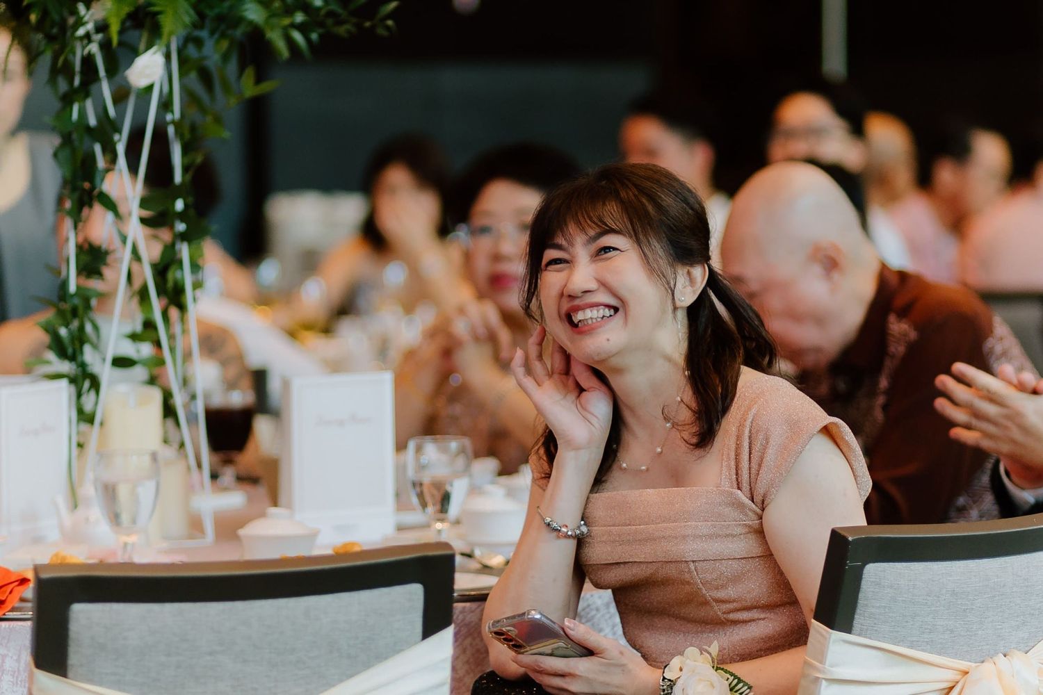 A woman smiles joyfully at a wedding reception dinner, seated at a decorated table with guests in the background.