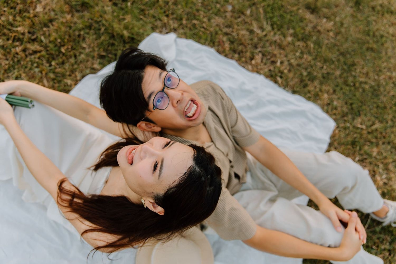 Two young women laughing and lying on a white blanket on grass, enjoying a sunny day outdoors together.