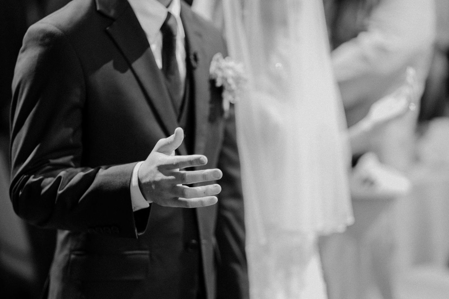 Black and white photo of a groom in a suit clasping hands with a bride in a veil at a wedding ceremony.