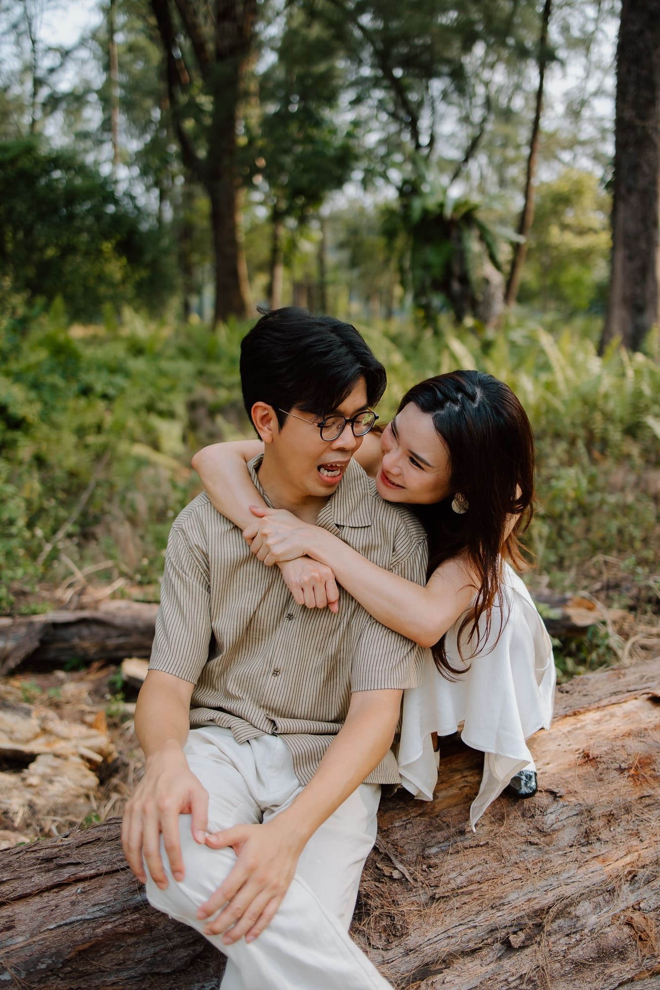 A young couple sitting on a log in a forest, the woman hugging the man from behind, both smiling warmly.