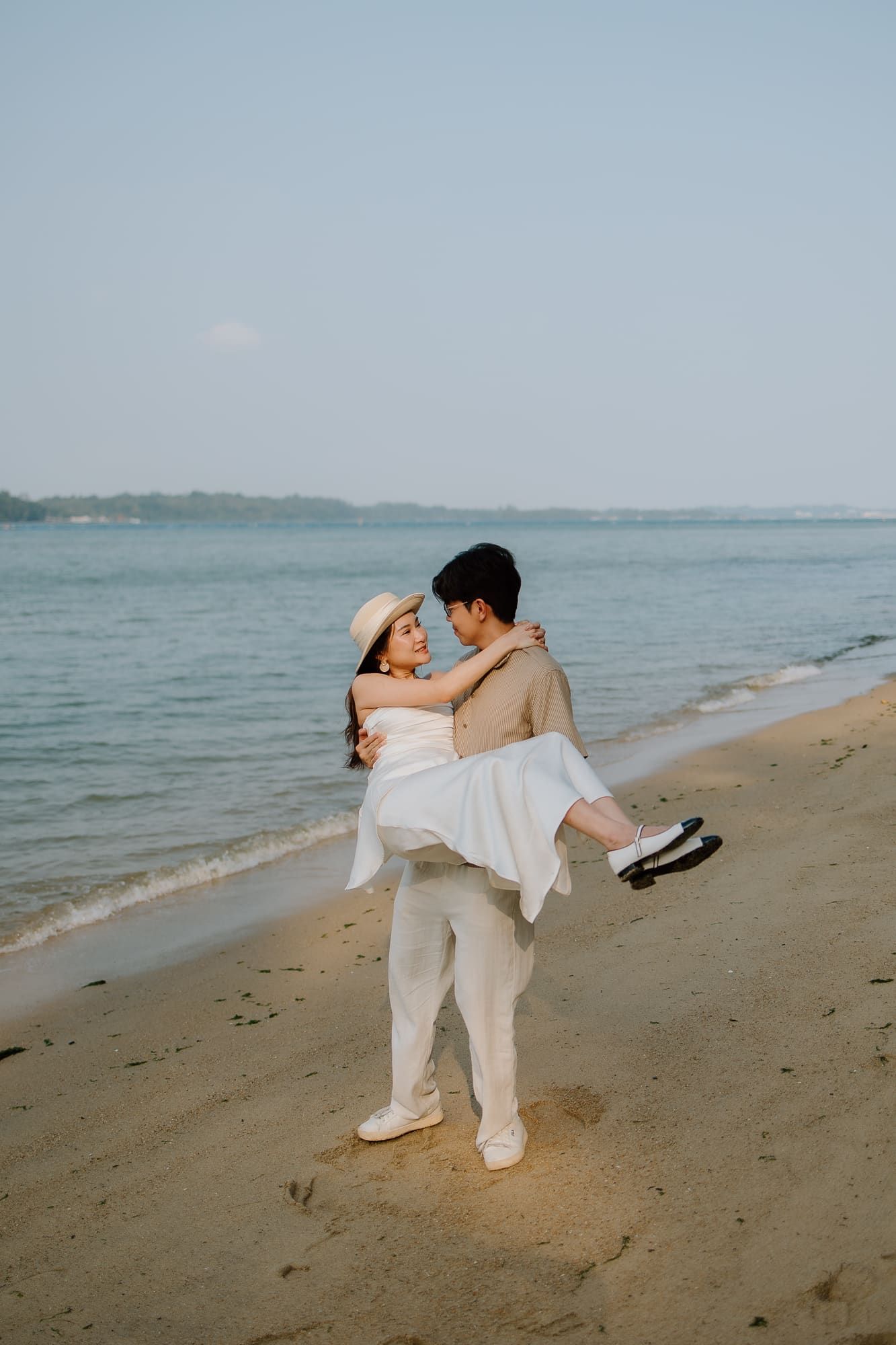 A groom carries his bride in a white dress and sun hat on a sandy beach with calm ocean waves behind them.