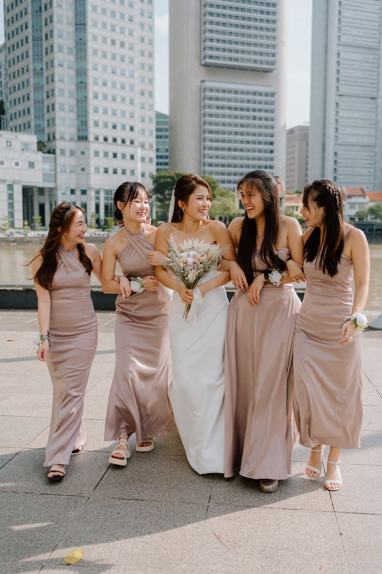A bride in white gown surrounded by bridesmaids in dusty rose dresses pose together against urban skyscrapers.