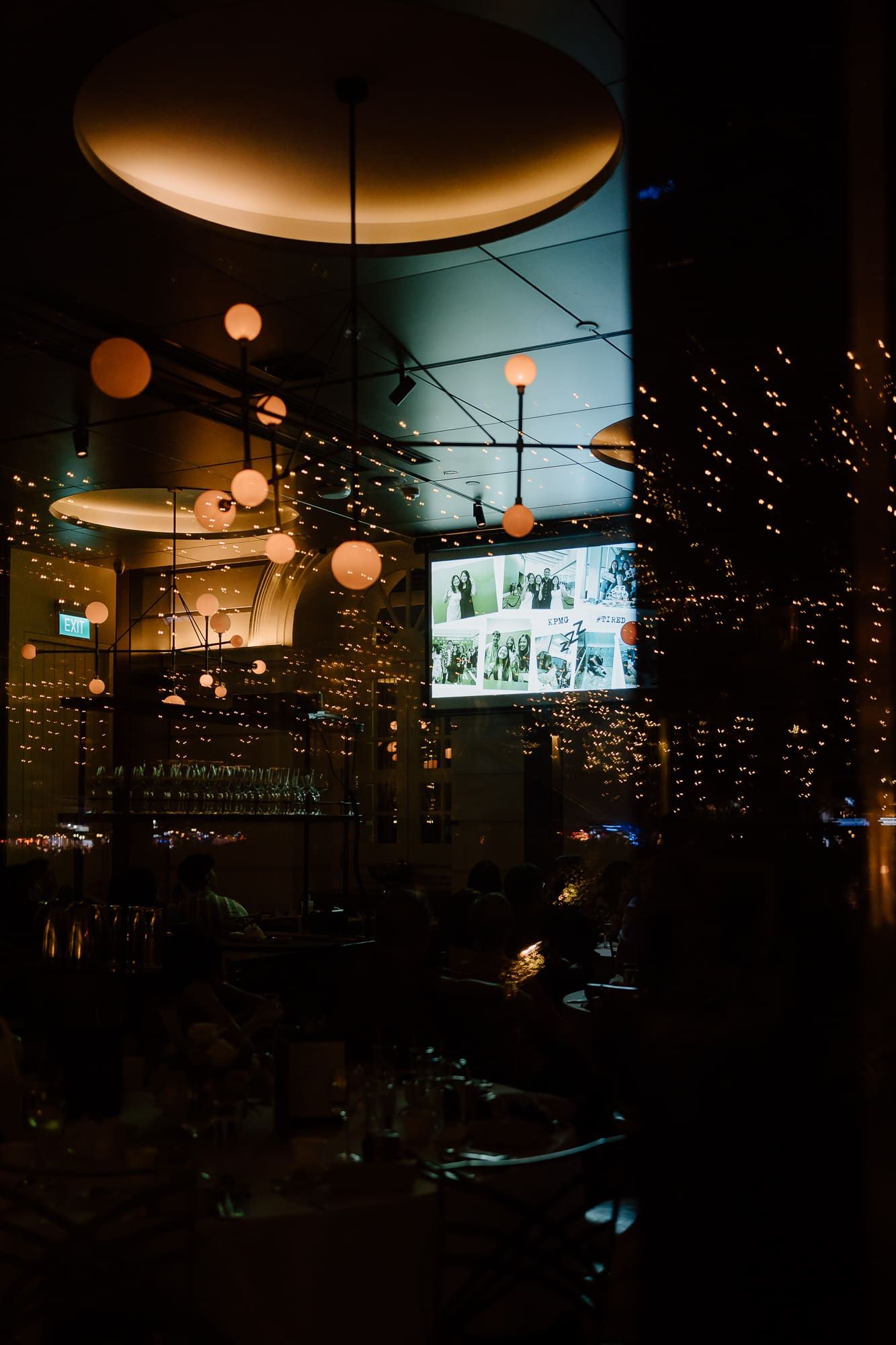 Dimly lit bar interior with moody ambiance featuring hanging pendant lights and a glowing wall display of bottles.