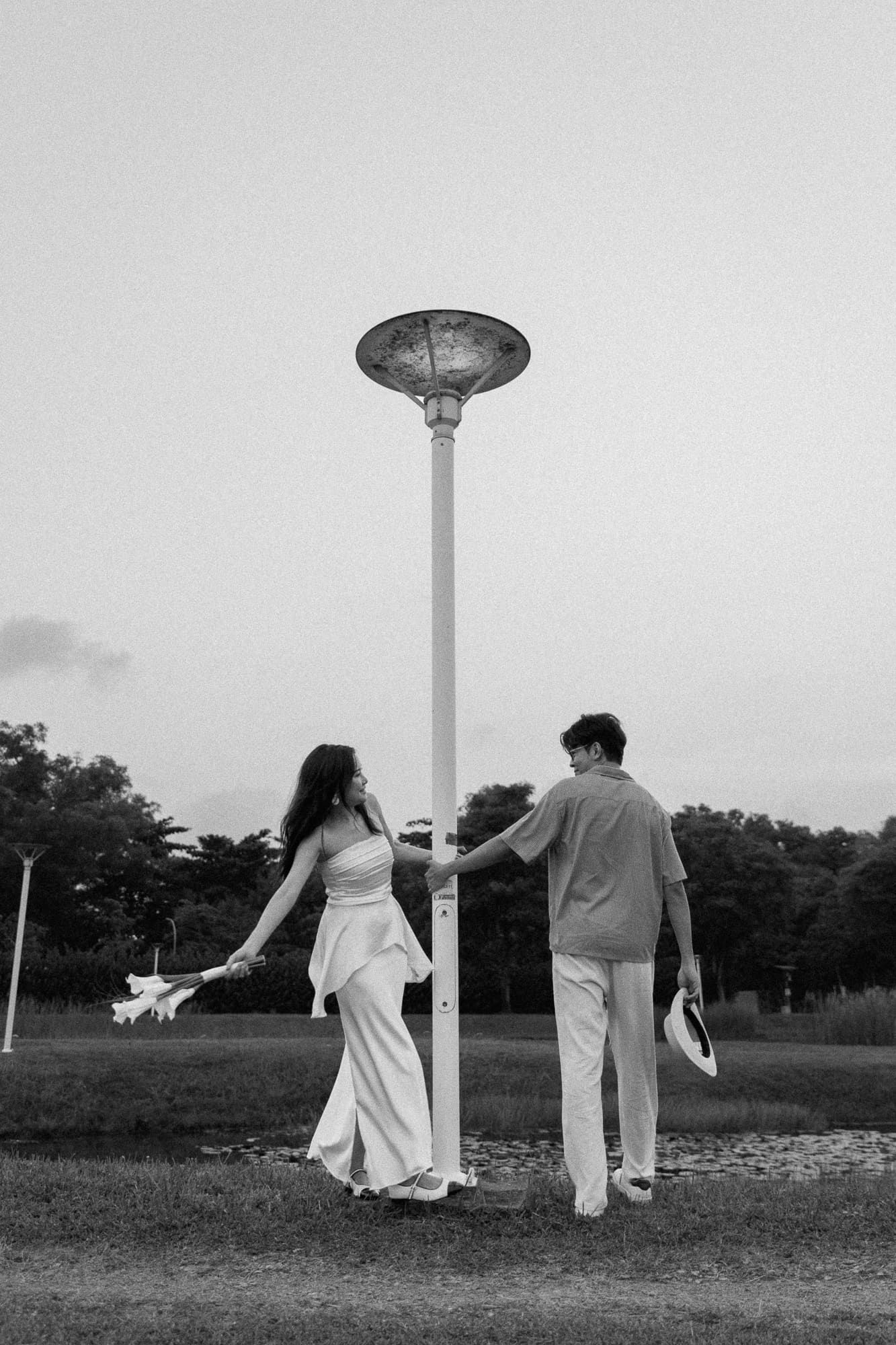 Black and white photo of a couple holding hands around a lamp post in a park at dusk.
