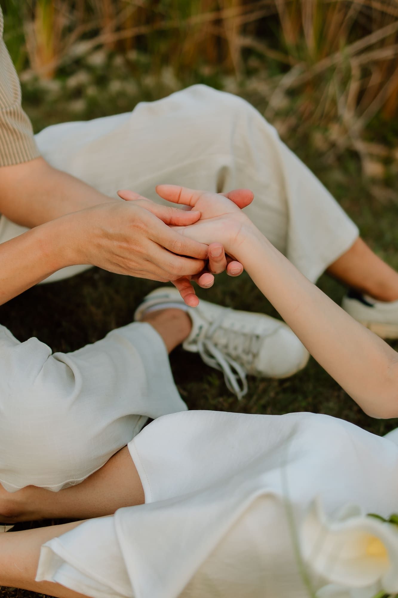 Two people sitting on grass holding hands tenderly, wearing white outfits, with a white flower nearby.