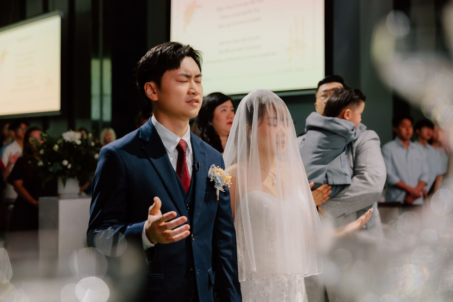 A groom in a navy suit and bride in a white wedding dress and veil stand at the altar during a church ceremony.