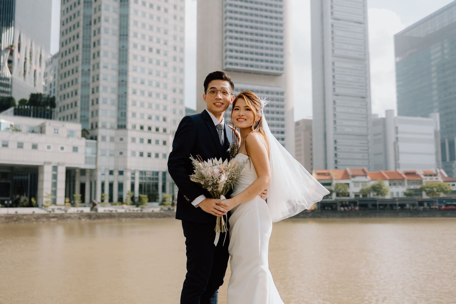 A couple poses in wedding attire along a waterfront with modern skyscrapers in the background.