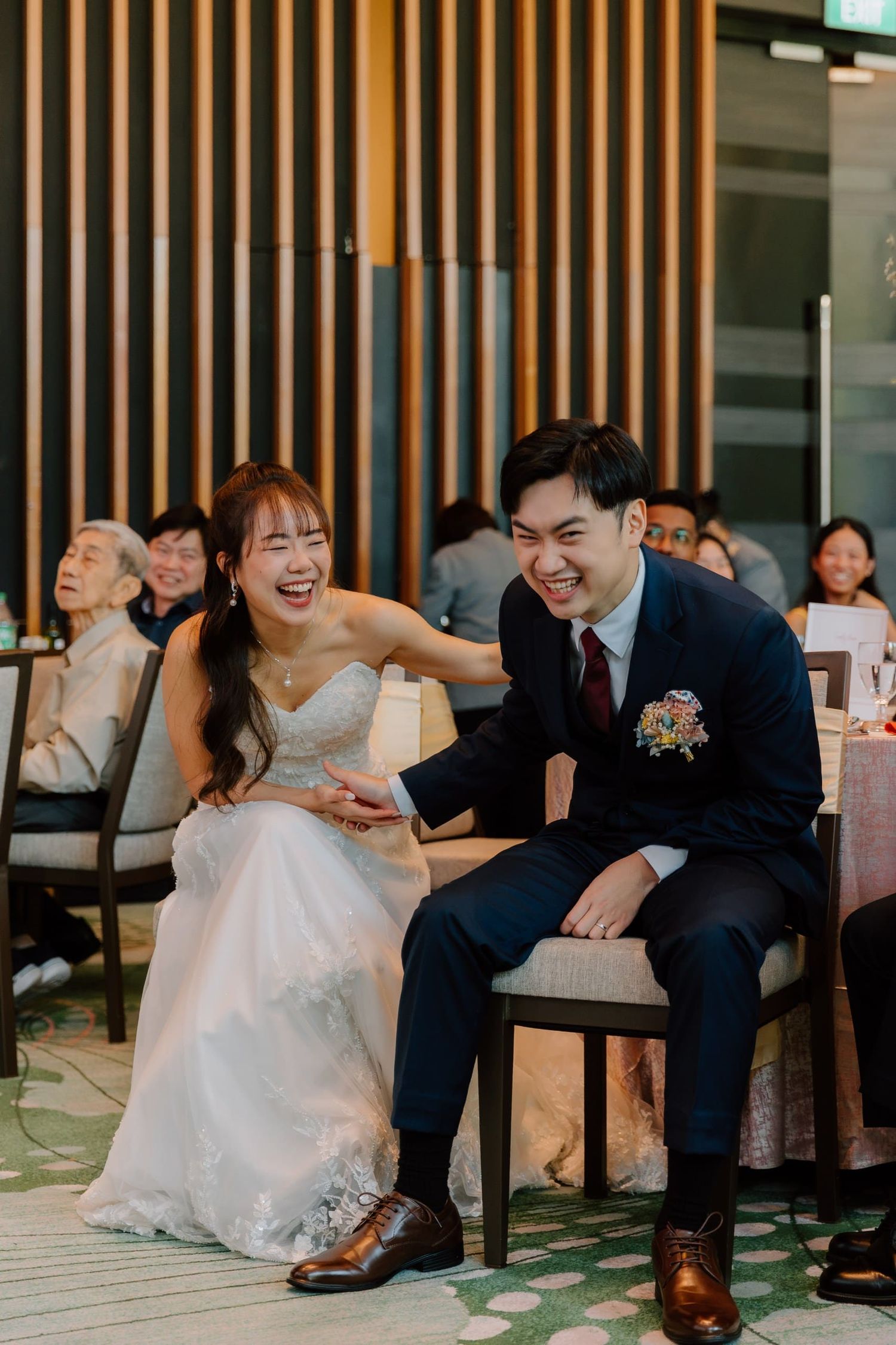 Bride and groom laughing joyfully together at their wedding reception, seated in elegant chairs at a banquet venue.