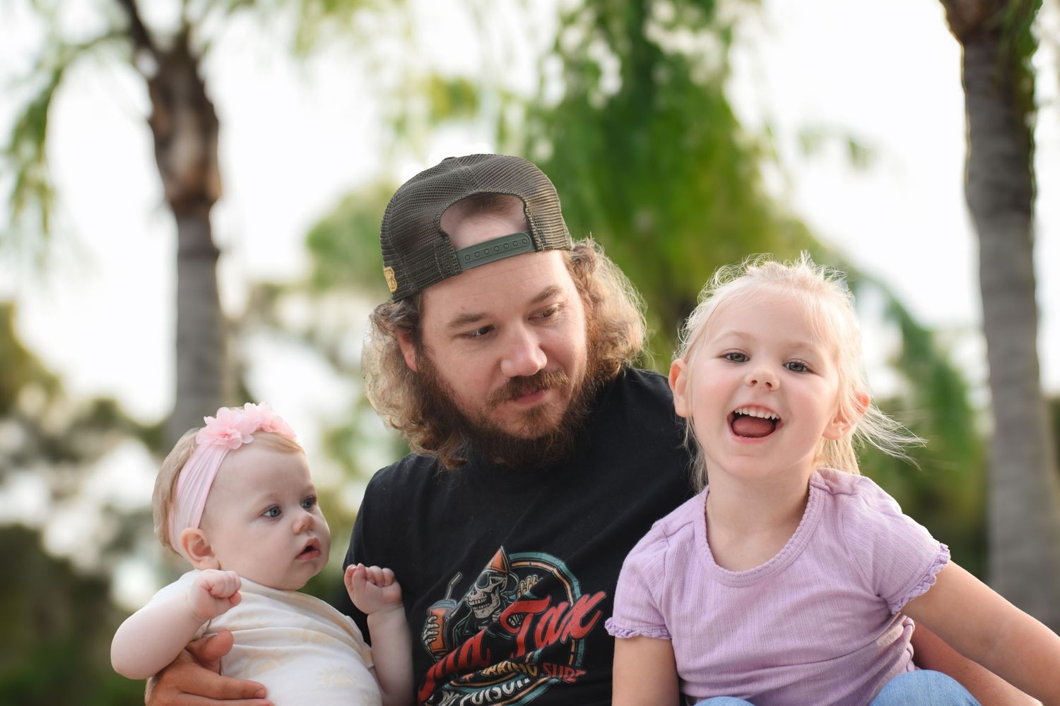 Happy bearded father holding infant and blonde toddler daughter smiling outdoors near palm trees.