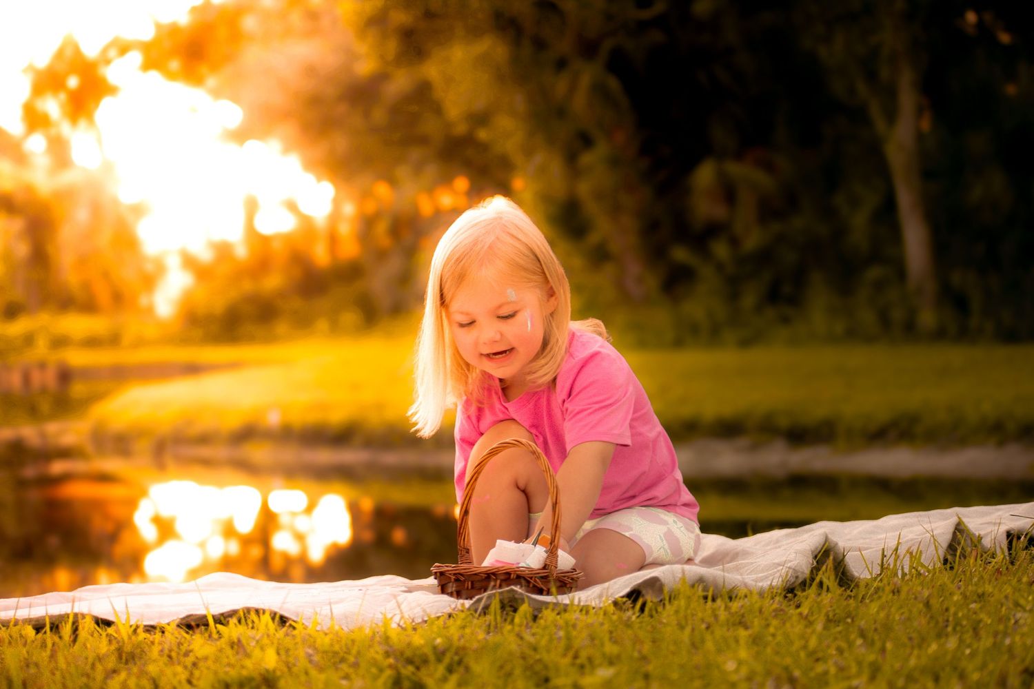 Toddler girl in pink shirt sitting on blanket by pond examining object at golden sunset.