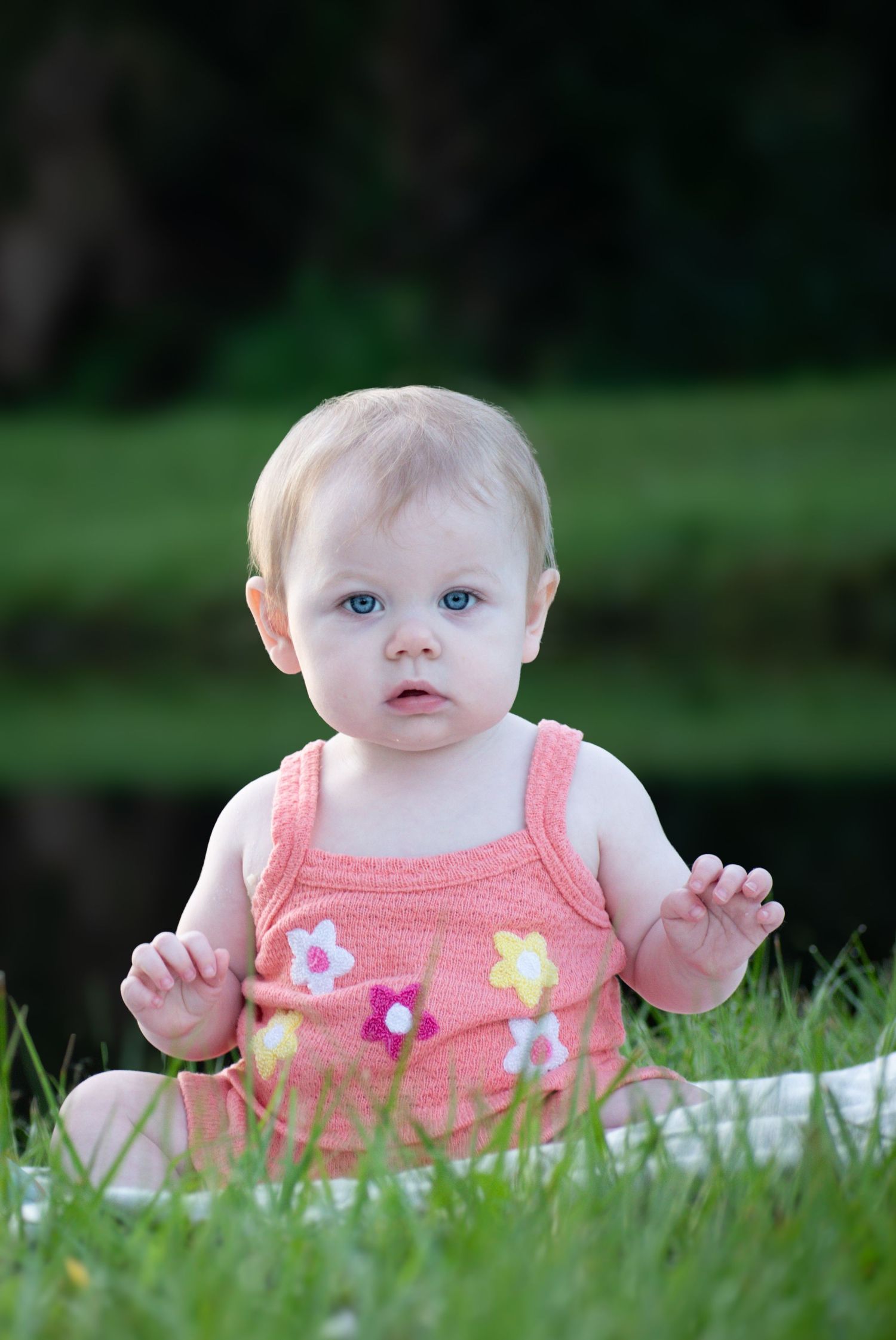 An adorable blue-eyed baby girl in a pink floral romper sits on grass outdoors.