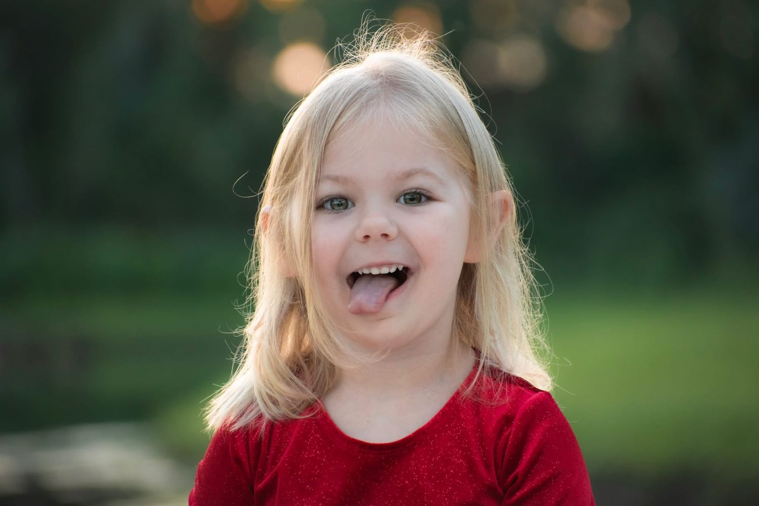 Playful blonde toddler girl in red shirt sticking out tongue with bokeh green background.