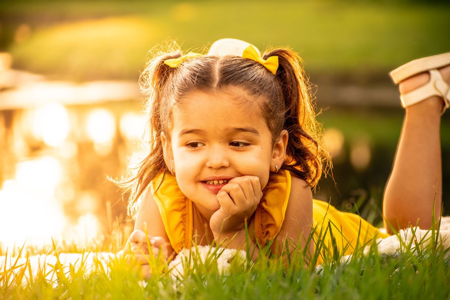 Cute little girl in yellow dress lying on grass smiling with golden sunset reflection behind.