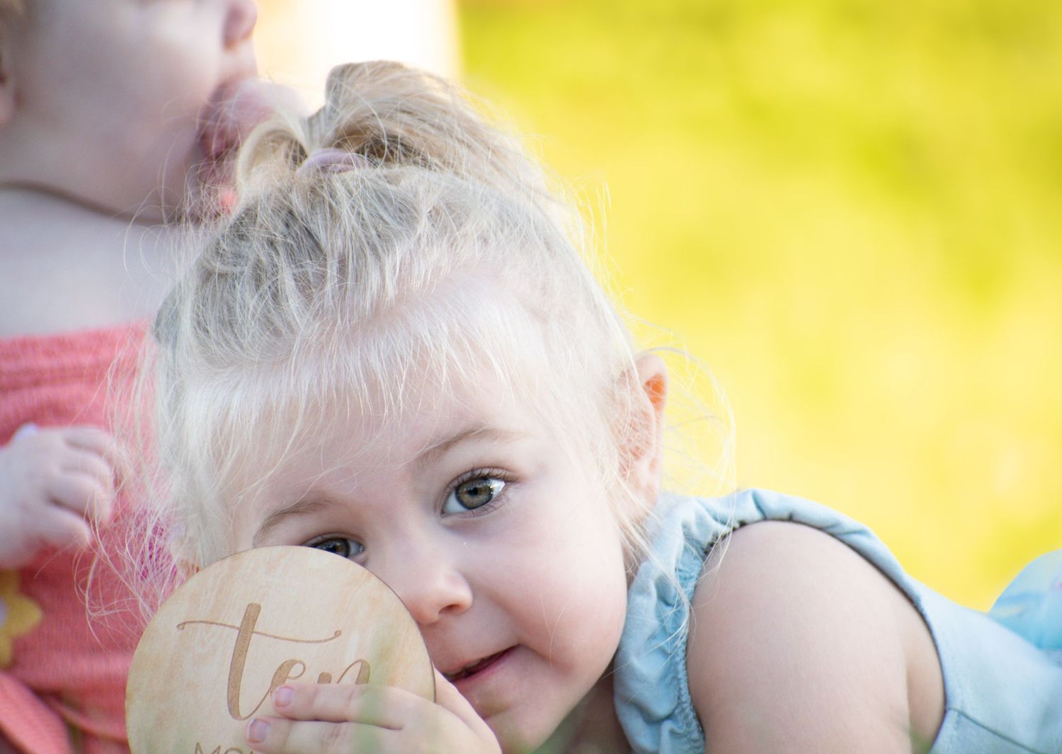 Close-up of blonde toddler girl in blue dress holding engraved wooden name disc outdoors in sunlight.
